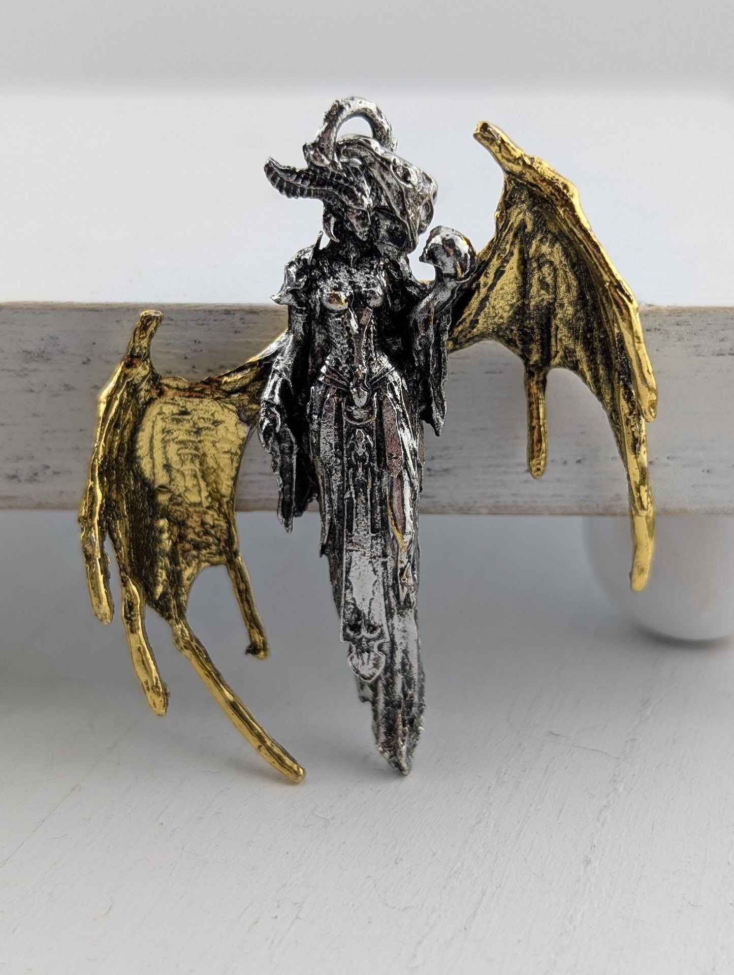 A pendant in the shape of a winged creature, possibly a dragon or mythical being, with intricate details and a metallic appearance.