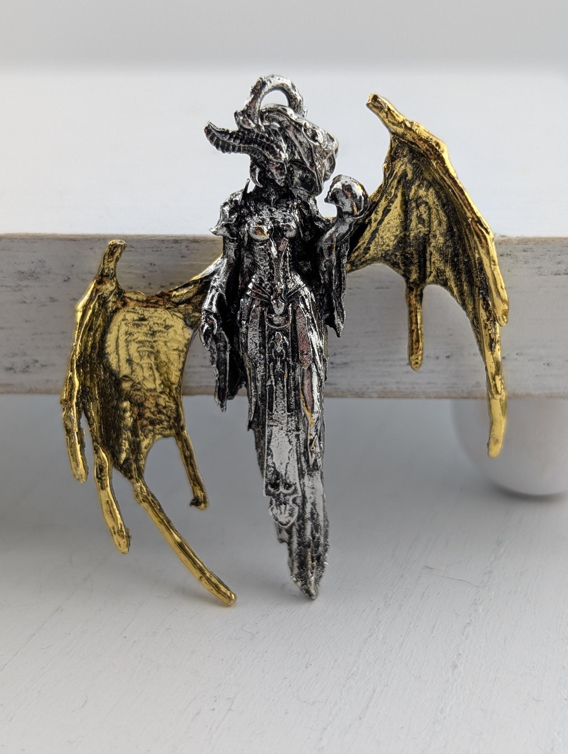 A pendant in the shape of a winged creature, possibly a dragon or mythical being, with intricate details and a metallic appearance.