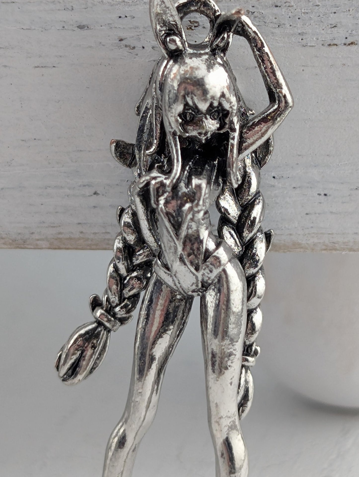 A silver-colored metal pendant in the shape of a female figure with long braided hair.