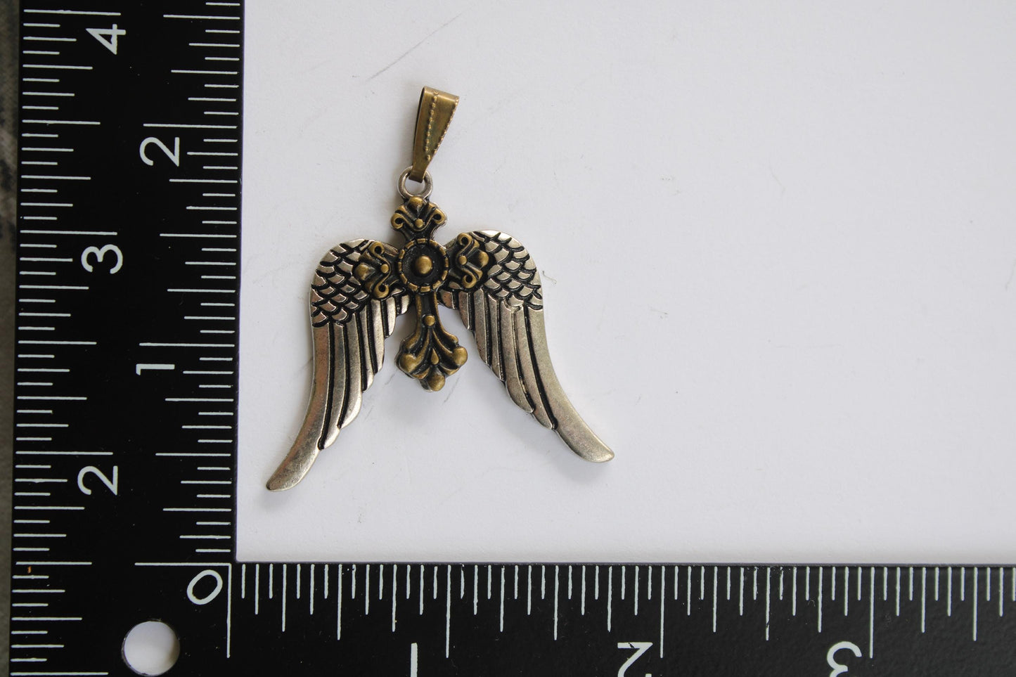 A pendant in the shape of a bird with wings, possibly a cross, is shown next to a ruler for size reference.