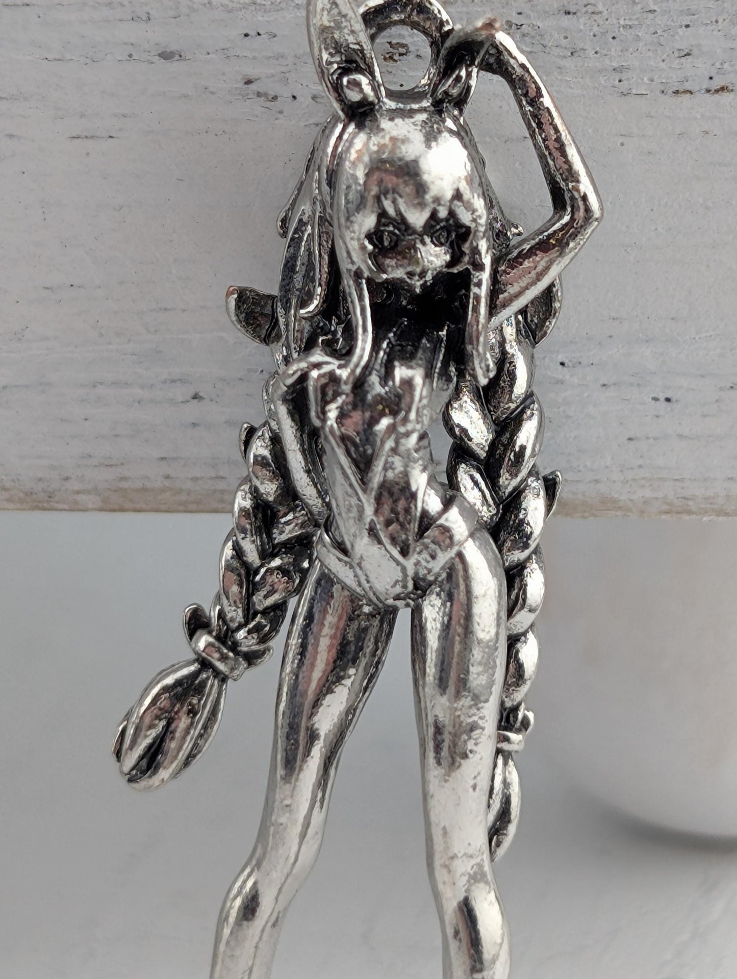 A silver-colored metal pendant in the shape of a female figure with long braided hair.