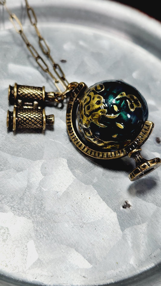 A decorative pendant with a globe design and a pair of binoculars hanging from it, resting on a metal surface.