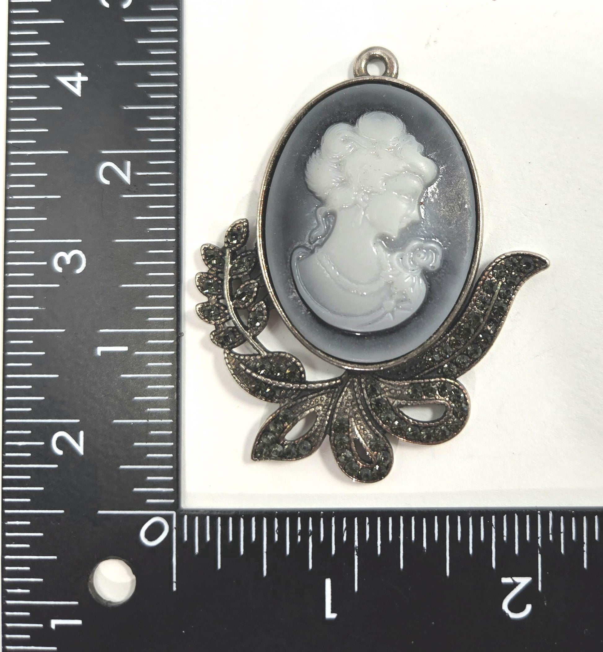 A pendant in the shape of a woman&#39;s profile, with a floral design and a metal frame. The pendant is placed next to a ruler for size comparison.