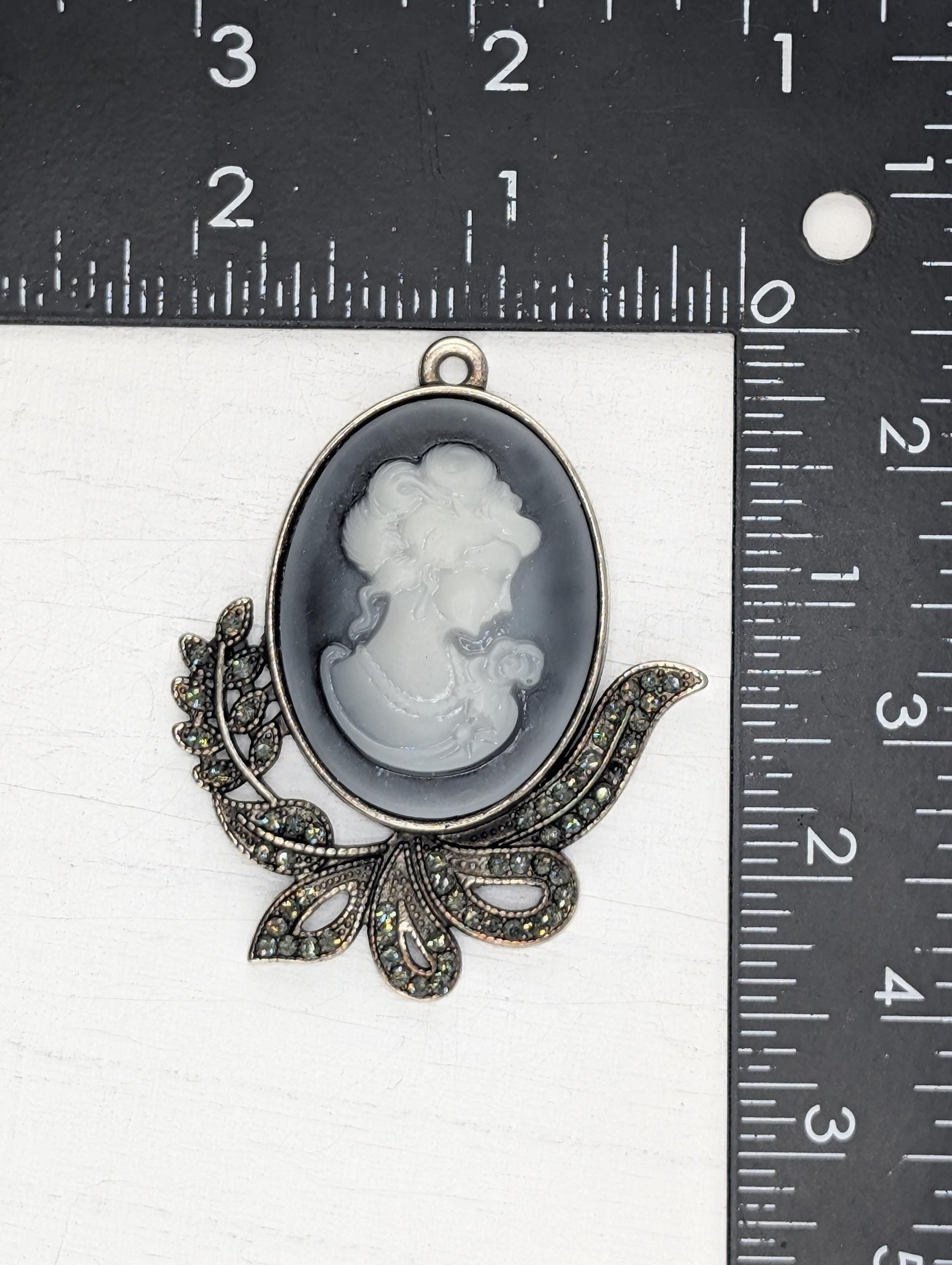 a pendant with a black and white oval-shaped design, possibly a profile portrait, and a decorative leaf-like element. The pendant is placed on a ruler for size comparison.