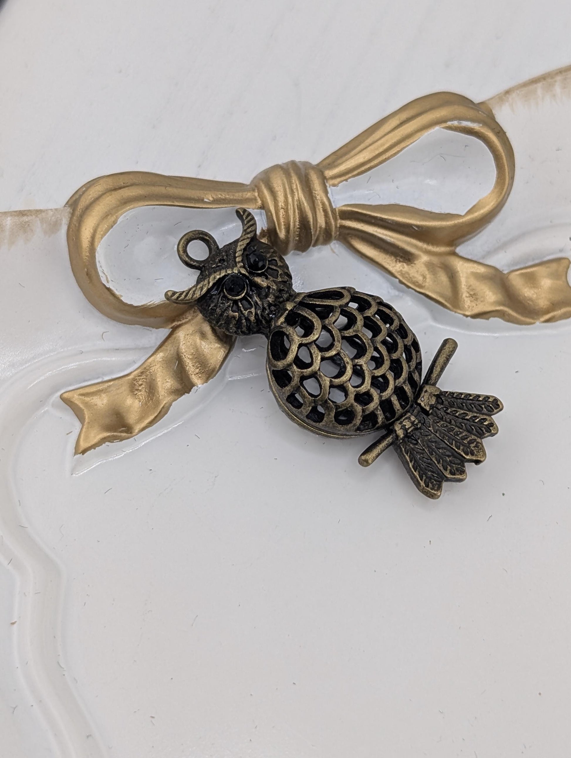 A decorative pendant featuring an owl charm is displayed on a white surface, with a gold ribbon bow.