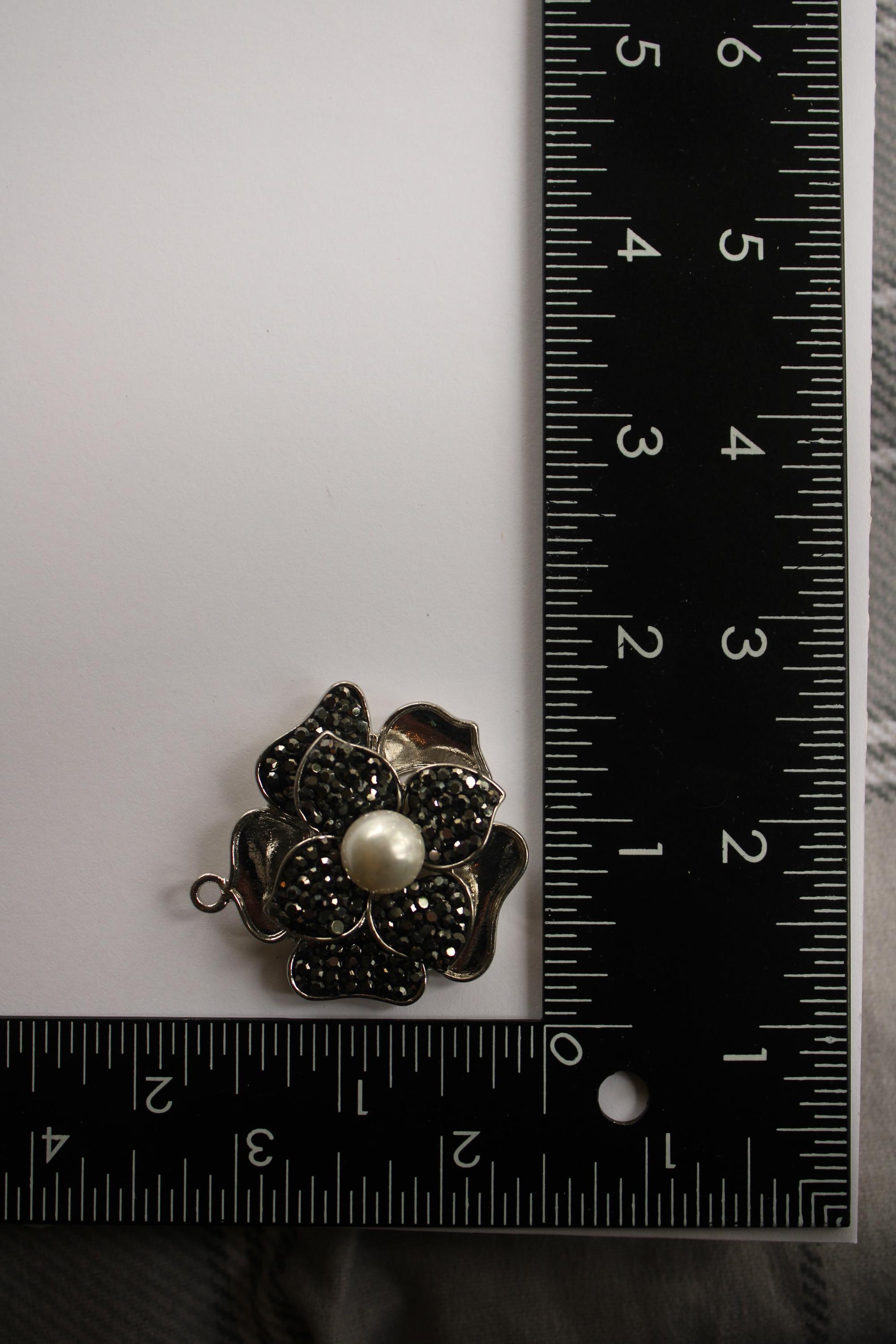A silver flower-shaped pendant with a pearl at its center is placed next to a ruler on a white surface.