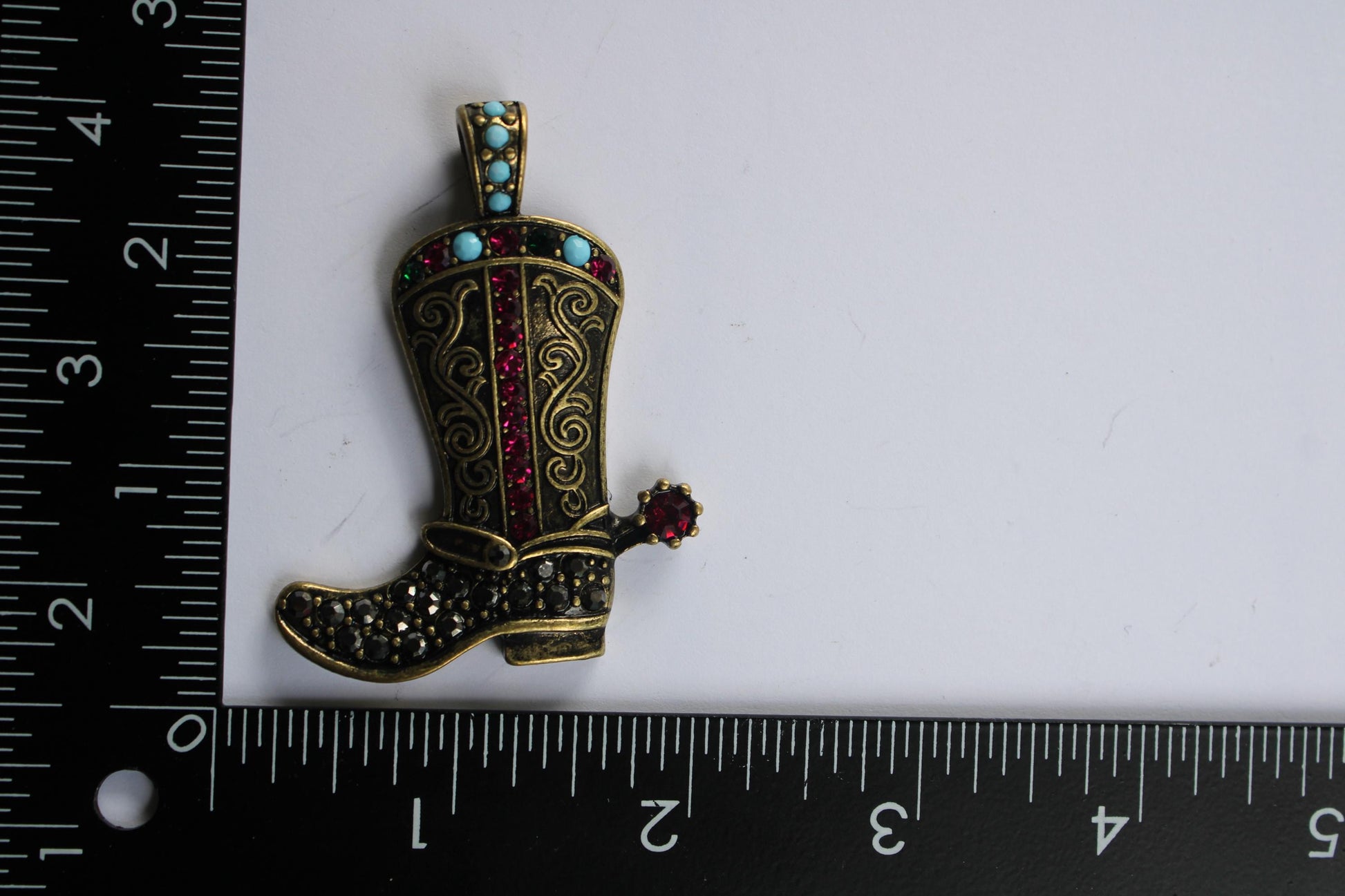 A decorative cowboy boot brooch with a red gemstone on the heel, placed next to a ruler for size reference.