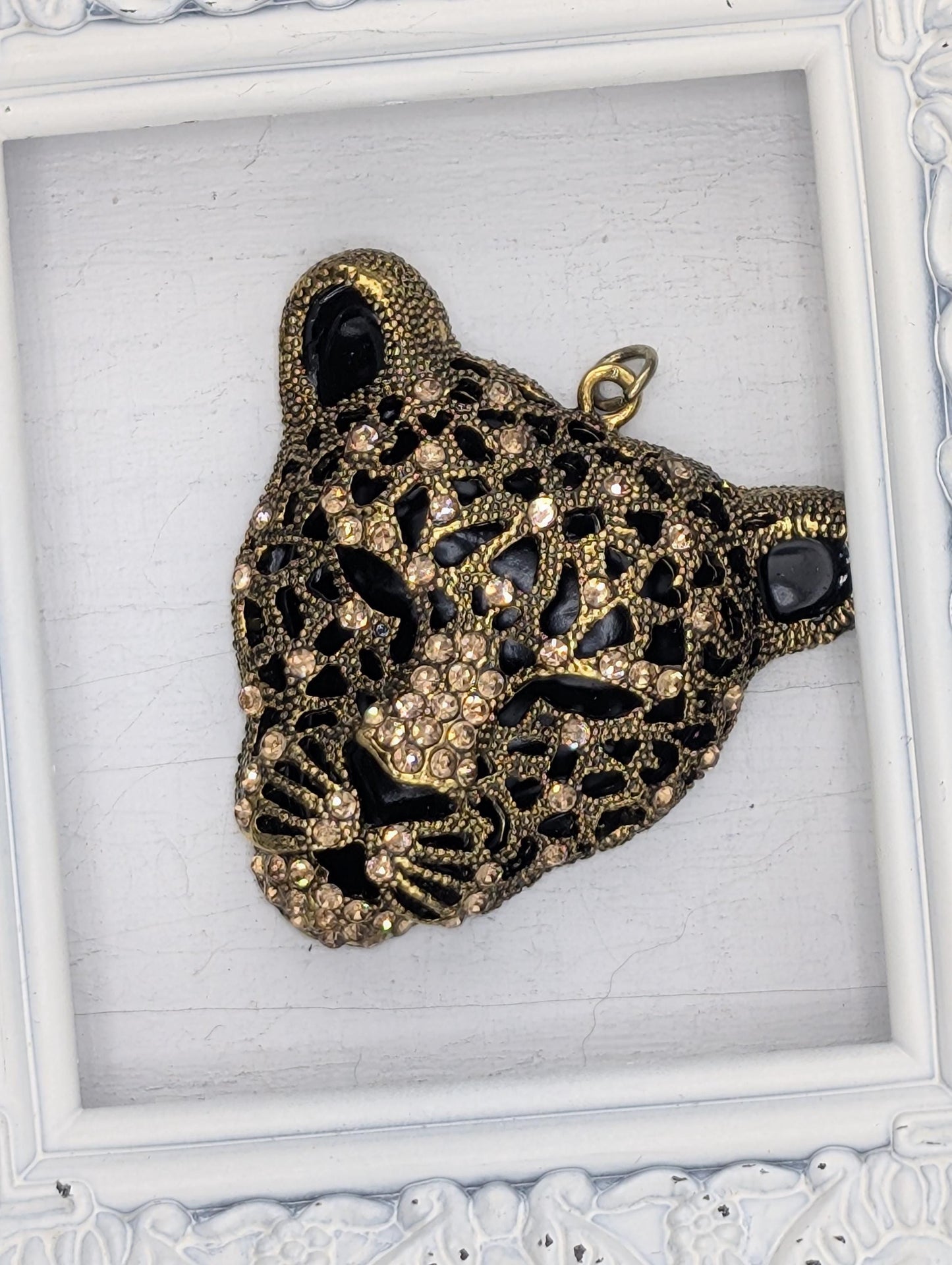 A decorative pendant in the shape of a leopard&#39;s head, adorned with intricate gold and black designs and sparkling rhinestones.