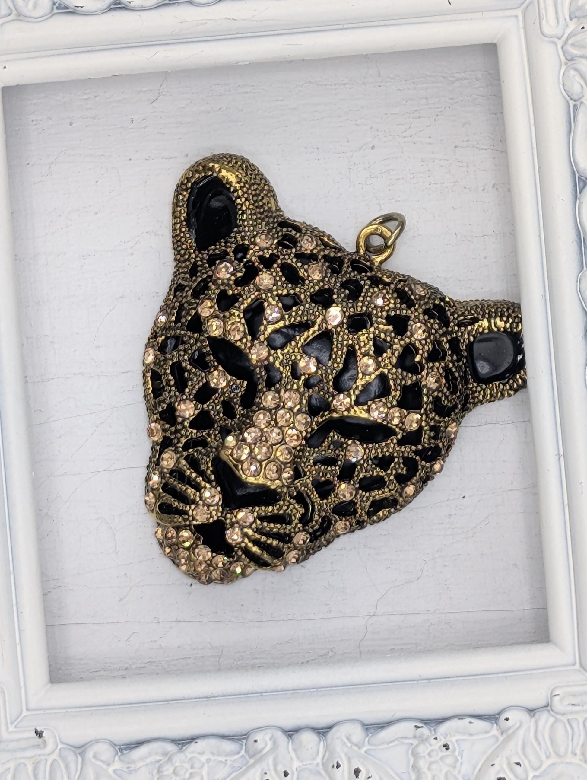 A decorative pendant in the shape of a leopard&#39;s head, adorned with intricate gold and black designs and sparkling rhinestones.
