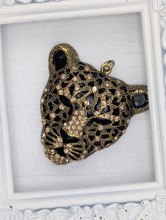 A decorative pendant in the shape of a leopard&#39;s head, adorned with intricate gold and black designs and sparkling rhinestones.