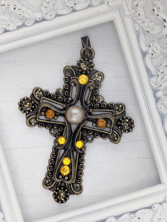 A decorative cross pendant with a pearl at the center, surrounded by intricate metalwork and adorned with gemstones.