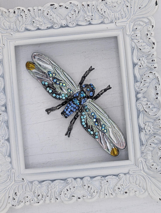 A decorative frame containing a large, intricately designed blue and silver insect, possibly a dragonfly or butterfly, with numerous small jewels or gemstones adorning its wings and body.