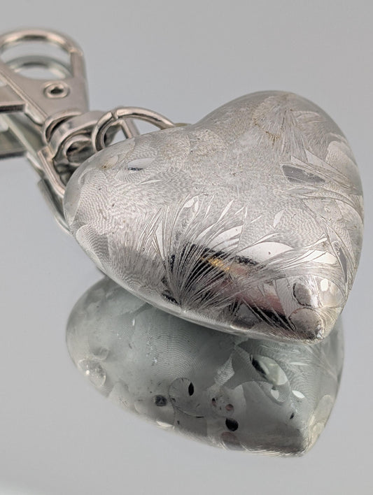 A silver heart-shaped keychain with an embossed floral pattern, resting on a reflective surface.