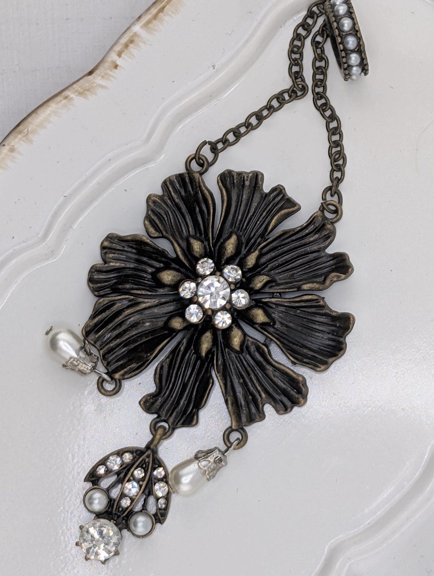 A decorative piece of jewelry featuring a large, intricately designed flower-shaped pendant with a central stone surrounded by smaller stones. The pendant is attached to a chain with pearls hanging from it, creating a stylish and elegant accessory.