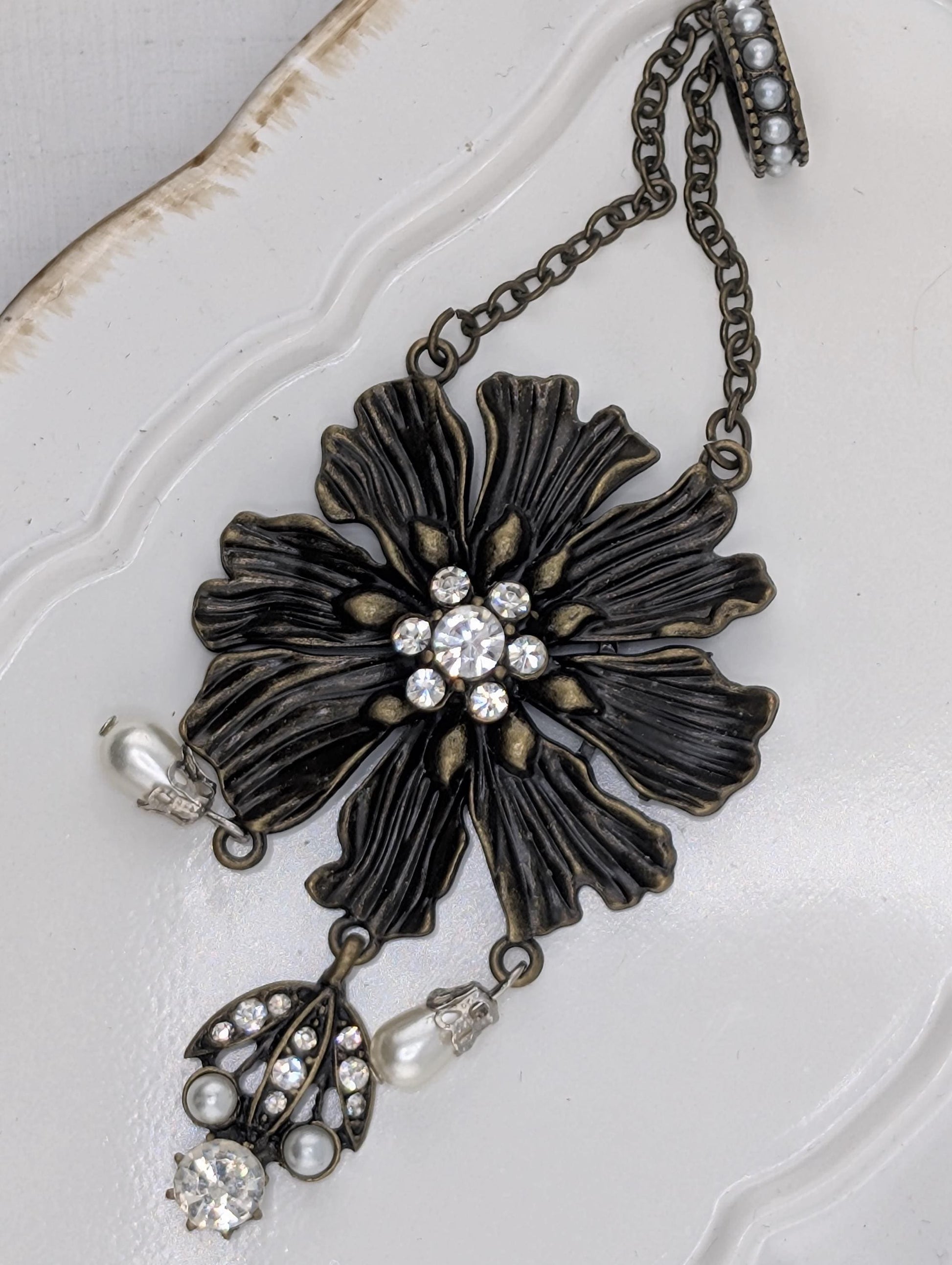 A decorative piece of jewelry featuring a large, intricately designed flower-shaped pendant with a central stone surrounded by smaller stones. The pendant is attached to a chain with pearls hanging from it, creating a stylish and elegant accessory.