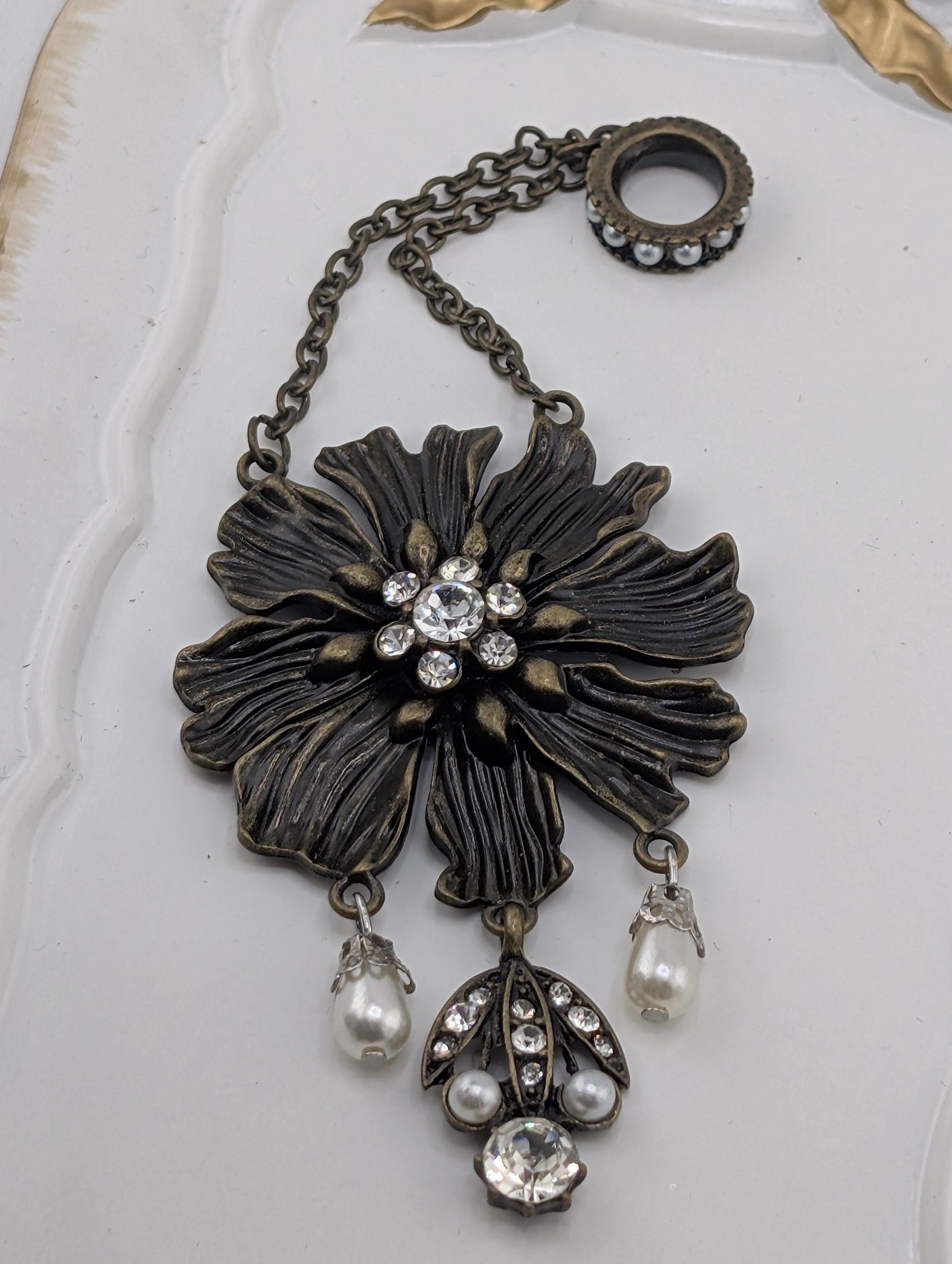 A decorative pendant featuring a large, intricately designed flower with a central stone surrounded by smaller stones, pearls, and dangling charms.