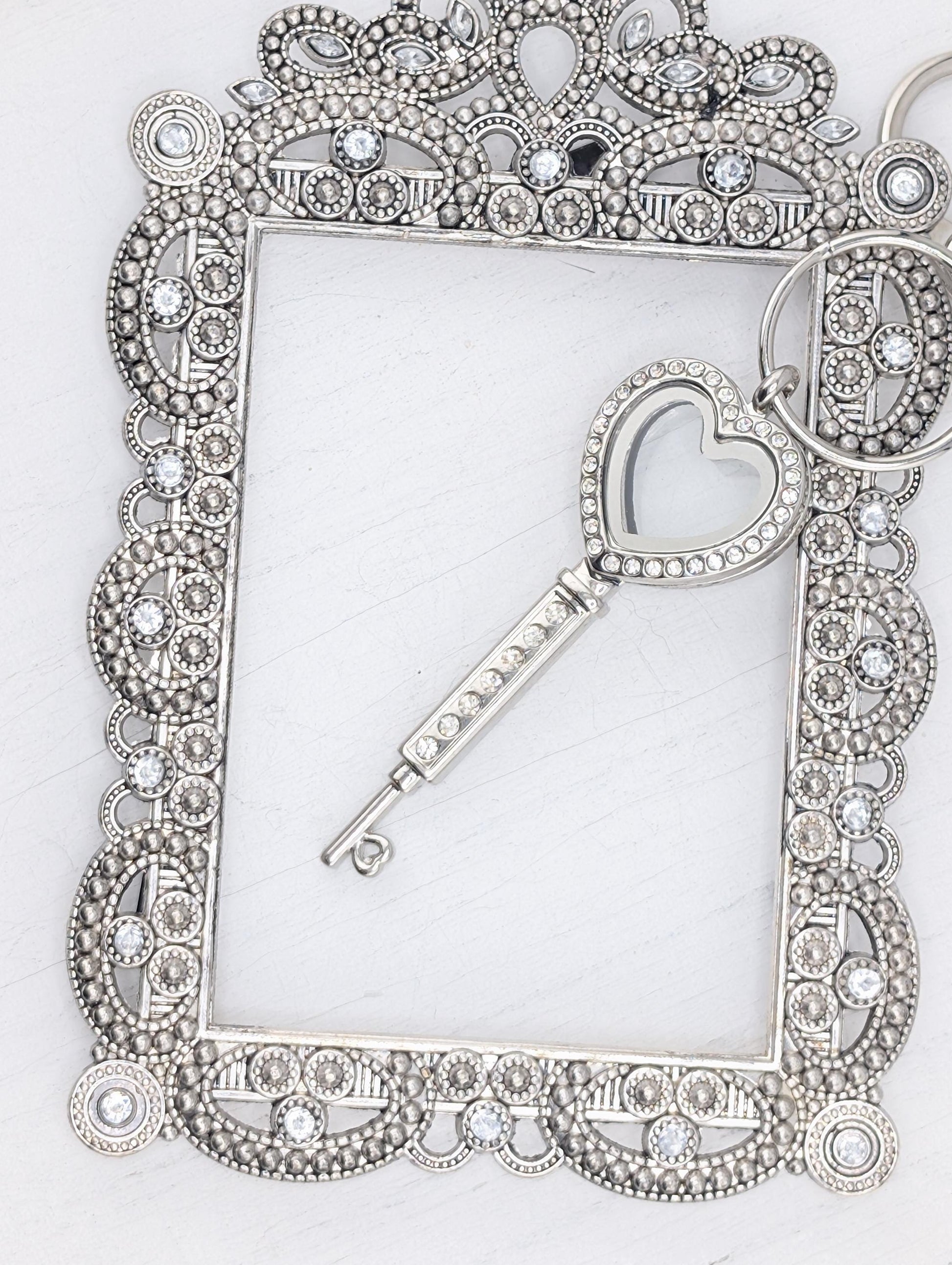 A silver-colored key with a heart-shaped cutout is placed within an ornate, decorative frame with a scalloped edge and a floral pattern.