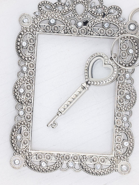 A silver-colored key with a heart-shaped cutout is placed within an ornate, decorative frame with a scalloped edge and a floral pattern.
