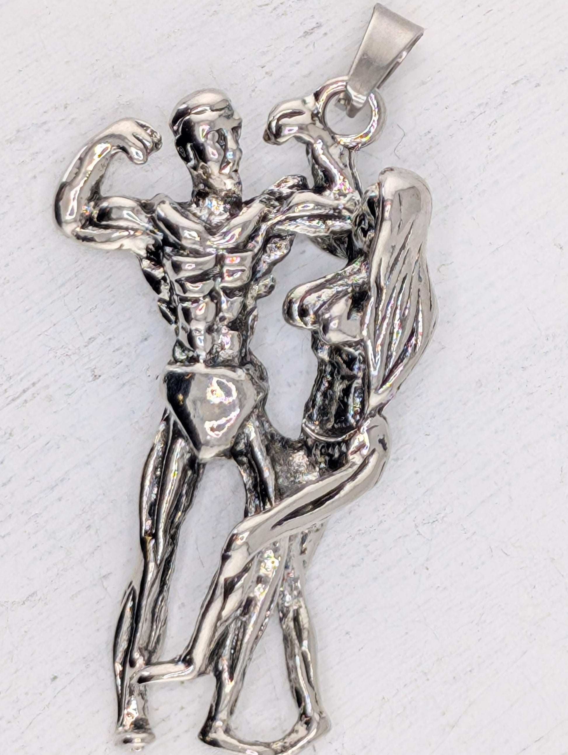 A silver-colored pendant in the shape of a muscular man and woman, with the man appearing to be flexing his muscles and the woman appearing to be in a dance-like pose.