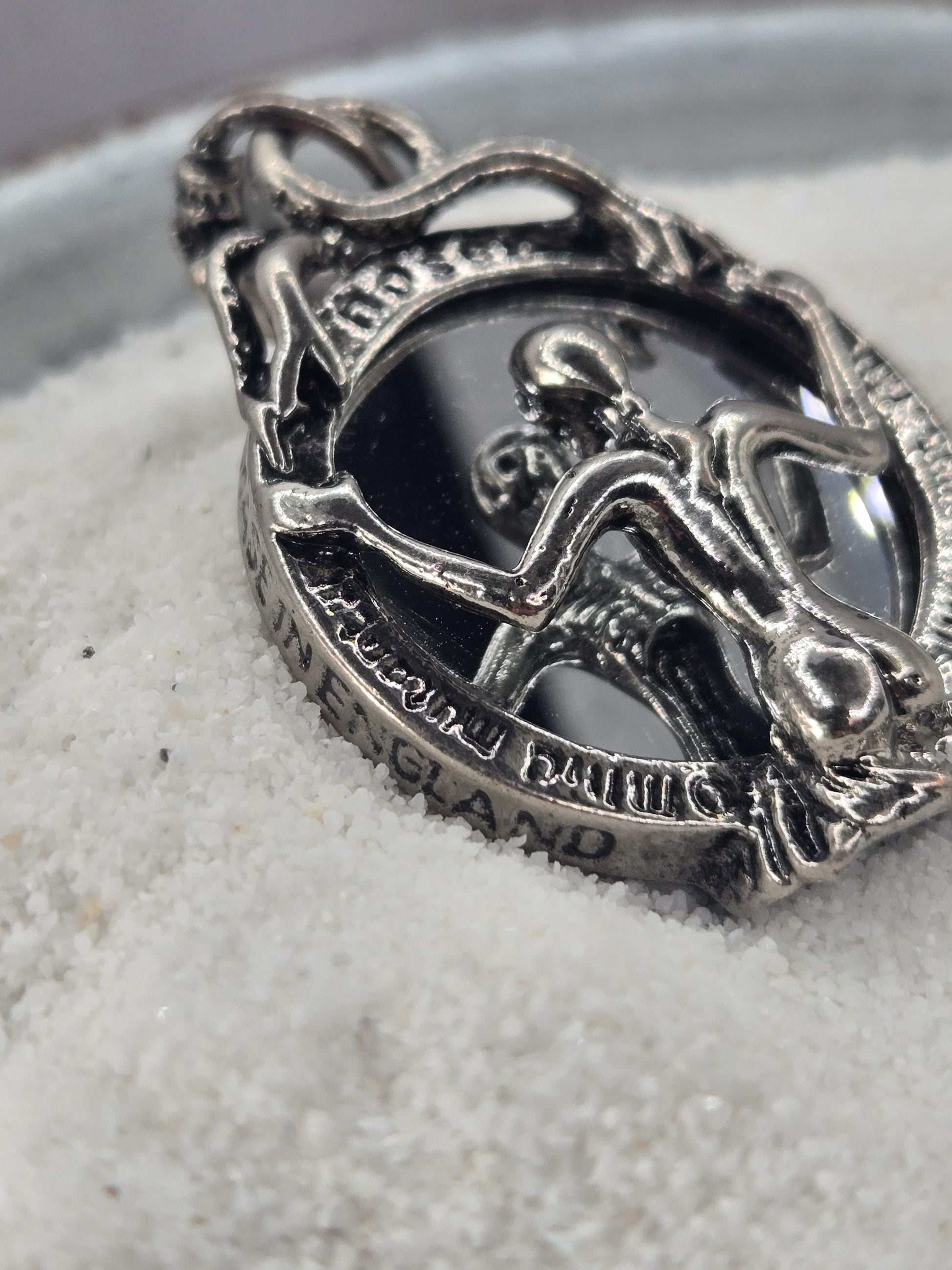 A close-up image of a silver-colored metal pendant or charm with an intricate design, resting on a white surface.