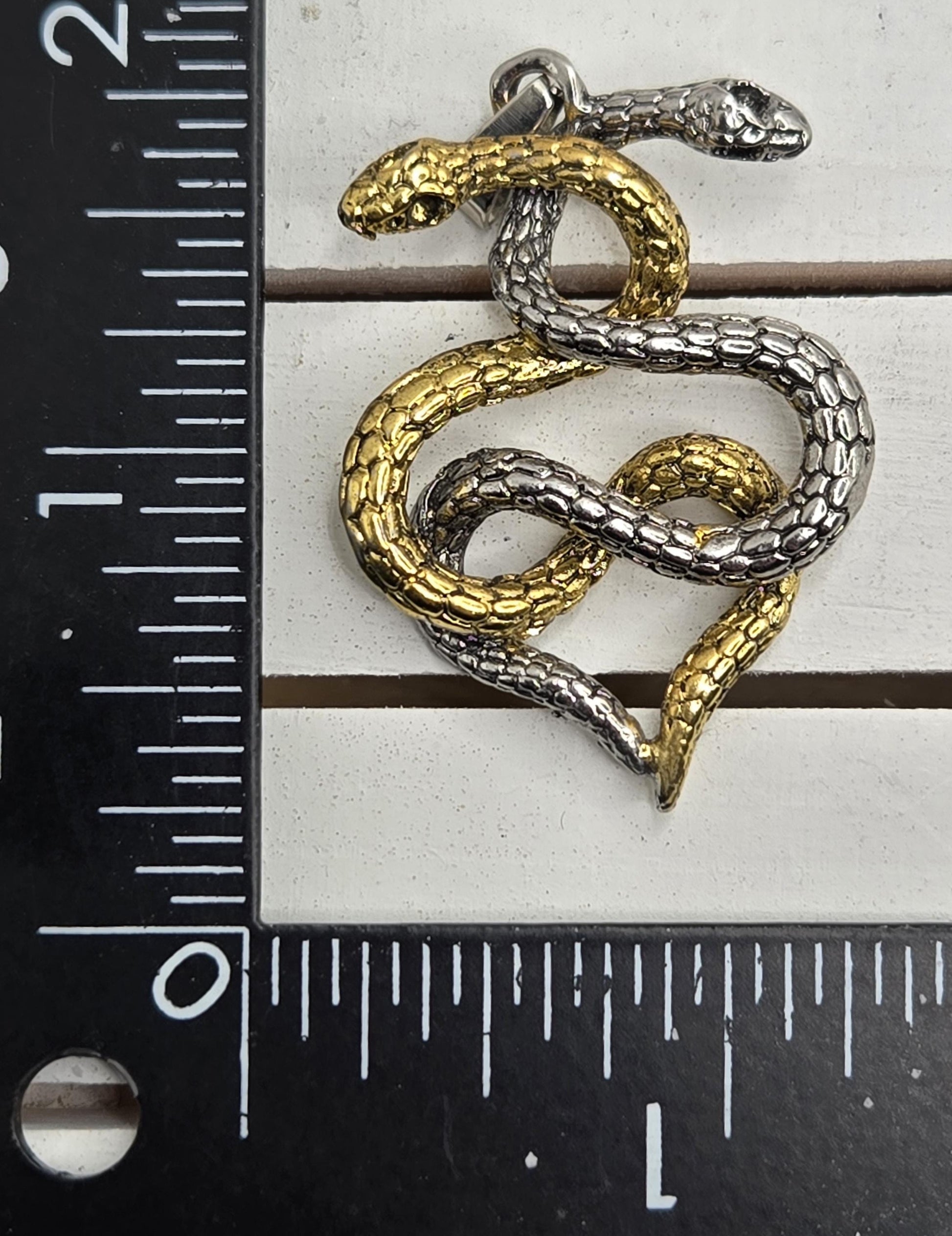 A close-up image of a pendant in the shape of a snake, with a gold and silver color scheme. The pendant is placed next to a ruler for size reference.