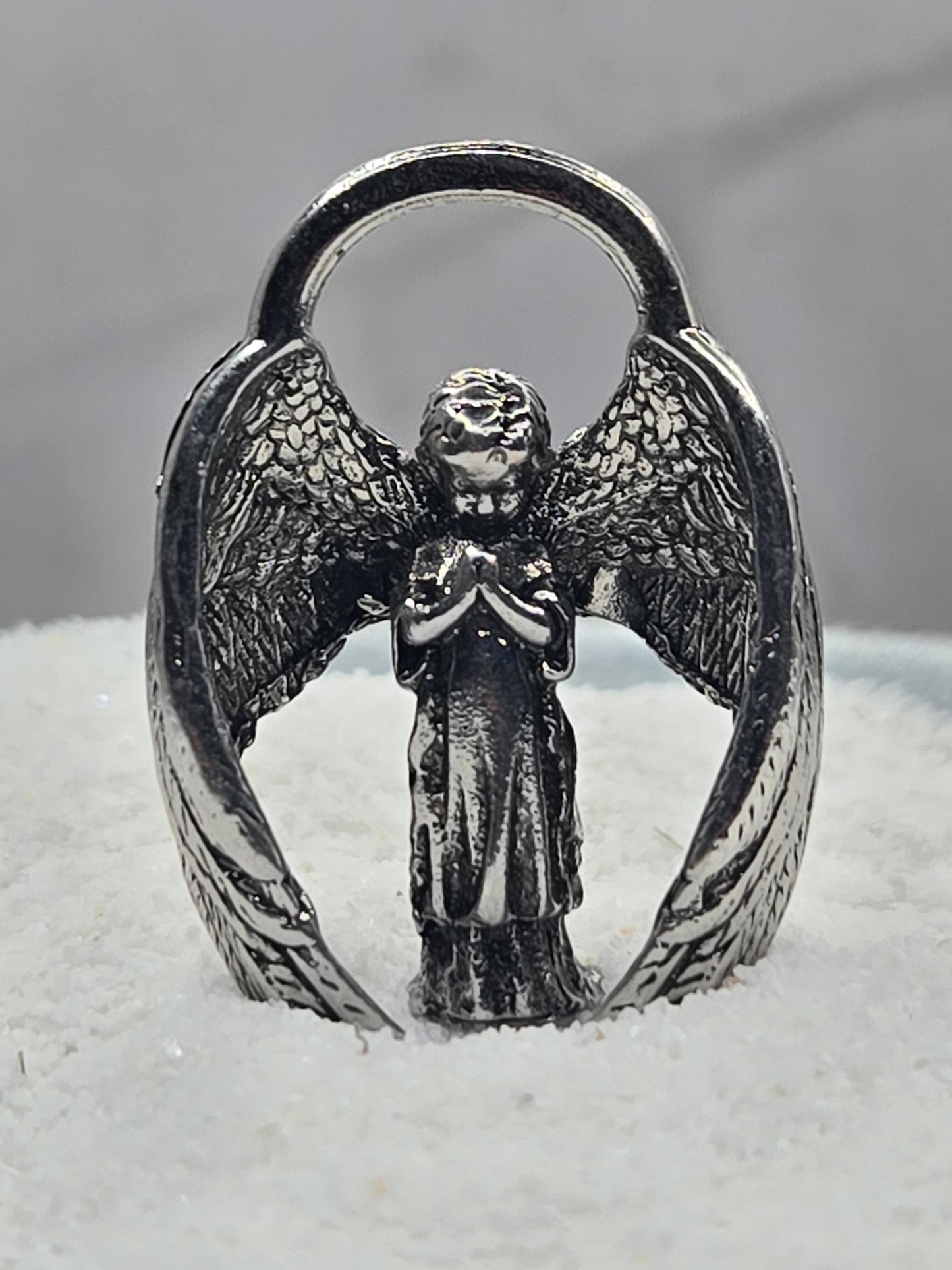 A silver-colored metal ring with an intricate design of wings and a central figure, possibly an angel or deity.
