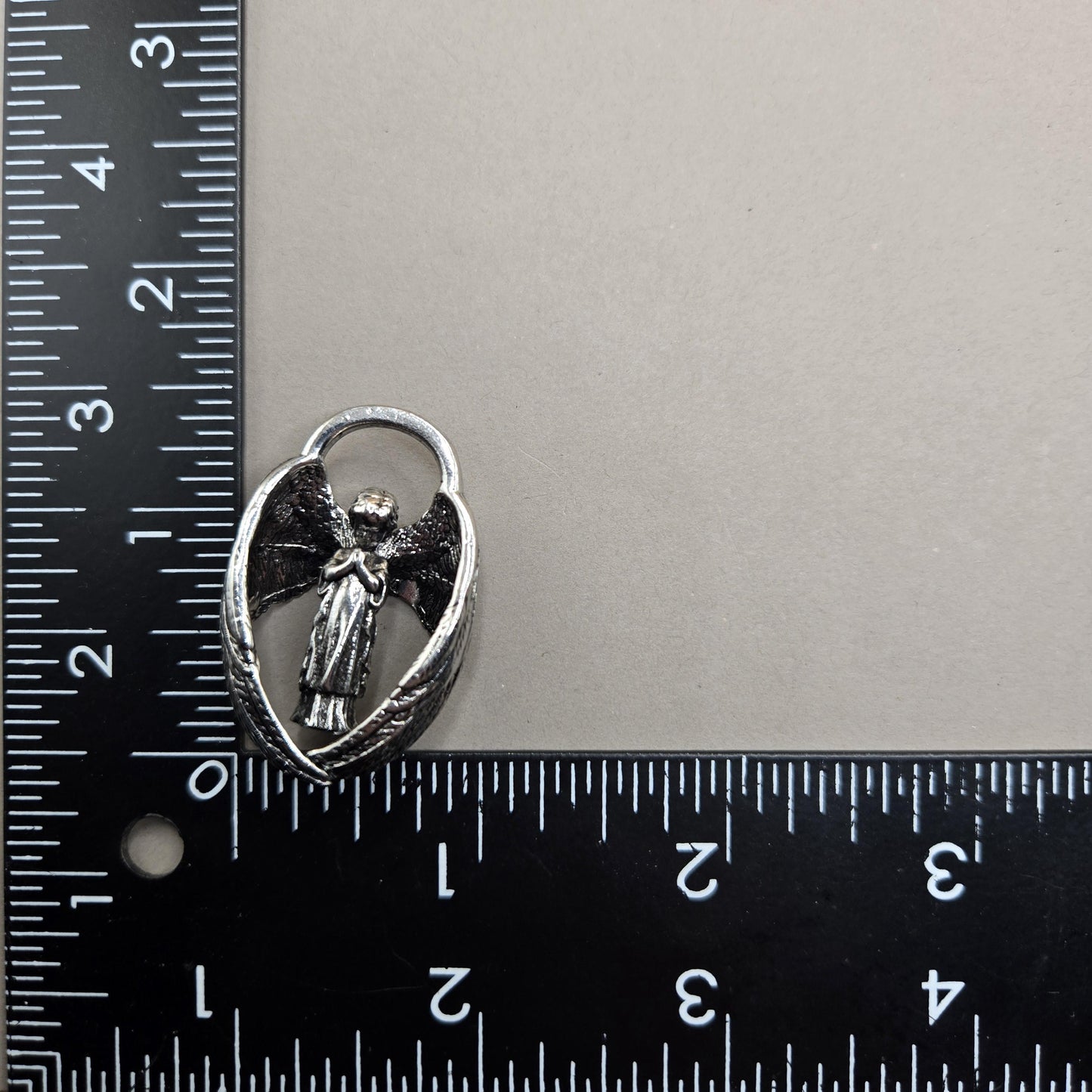 A silver-colored pendant or charm is placed next to a ruler on a gray surface.