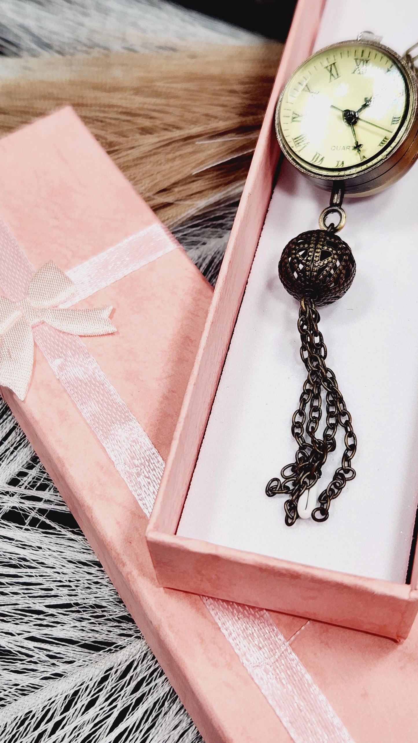 A pink box with a white ribbon, a small clock, and a chain with a ball on top, all placed on a pink surface.