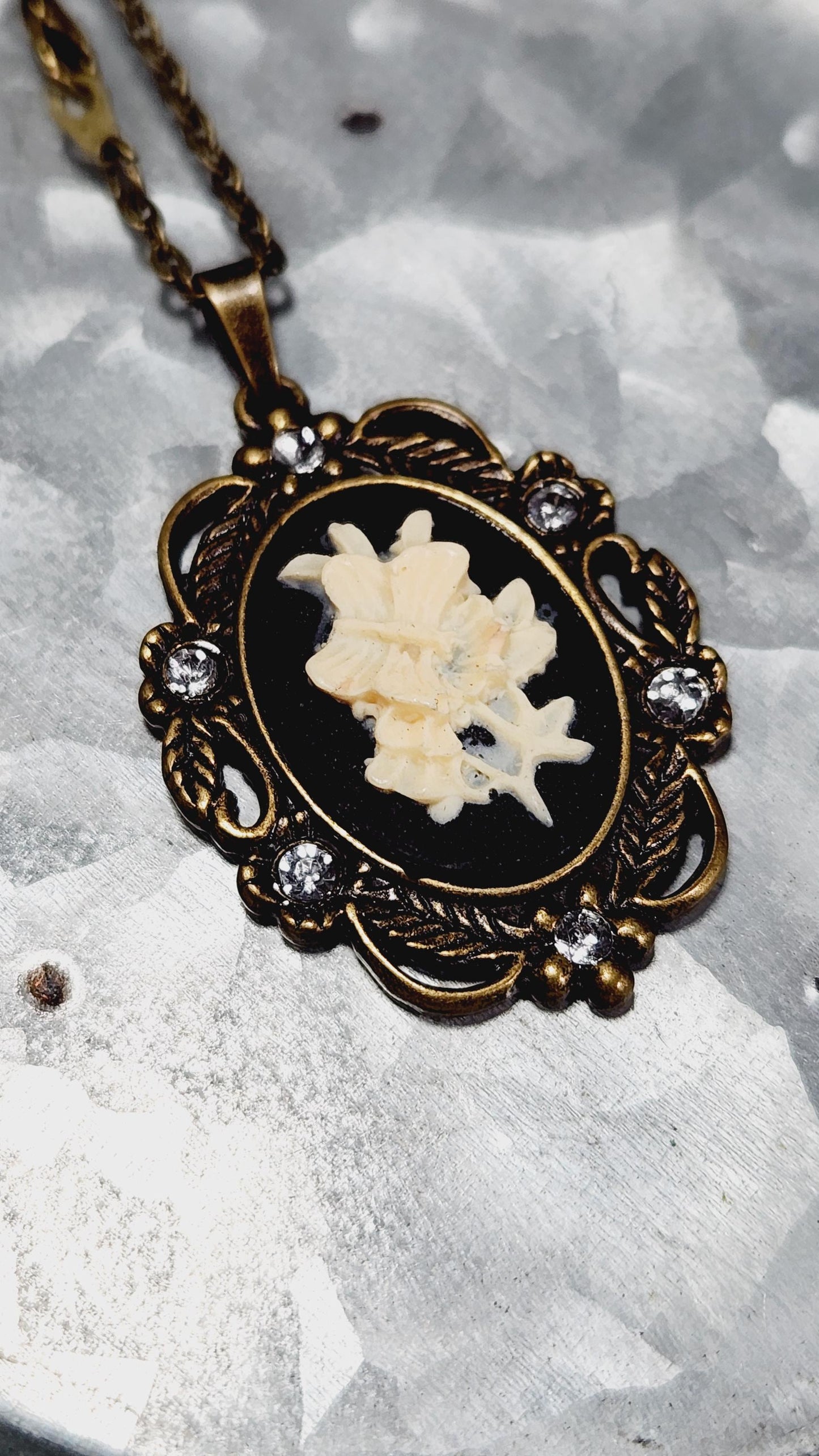 A decorative pendant with a floral design and small white stones is displayed on a chain against a textured gray background.
