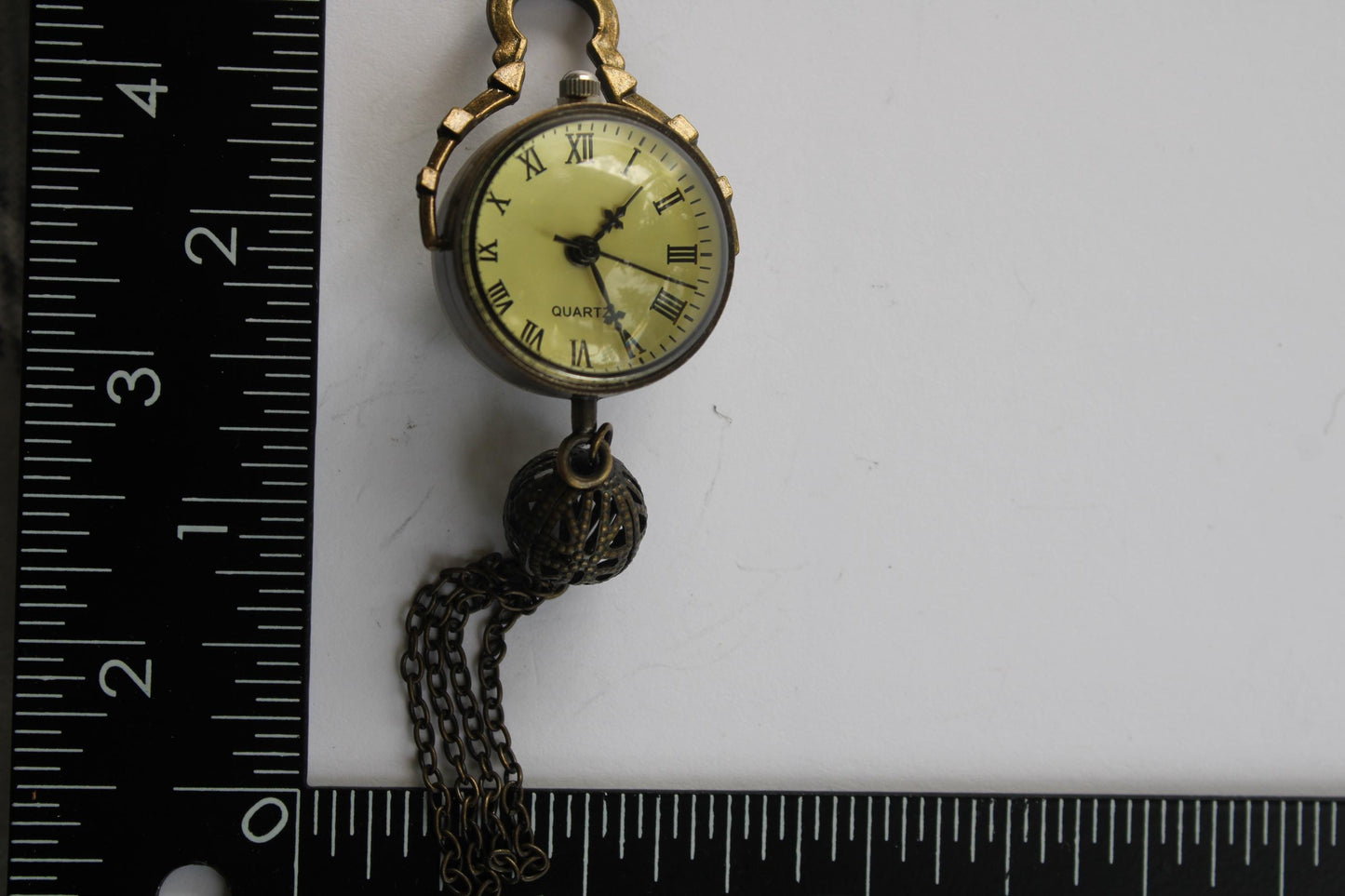 A small, round, gold-colored pocket watch with a tassel hanging from the chain is placed next to a ruler on a white surface.