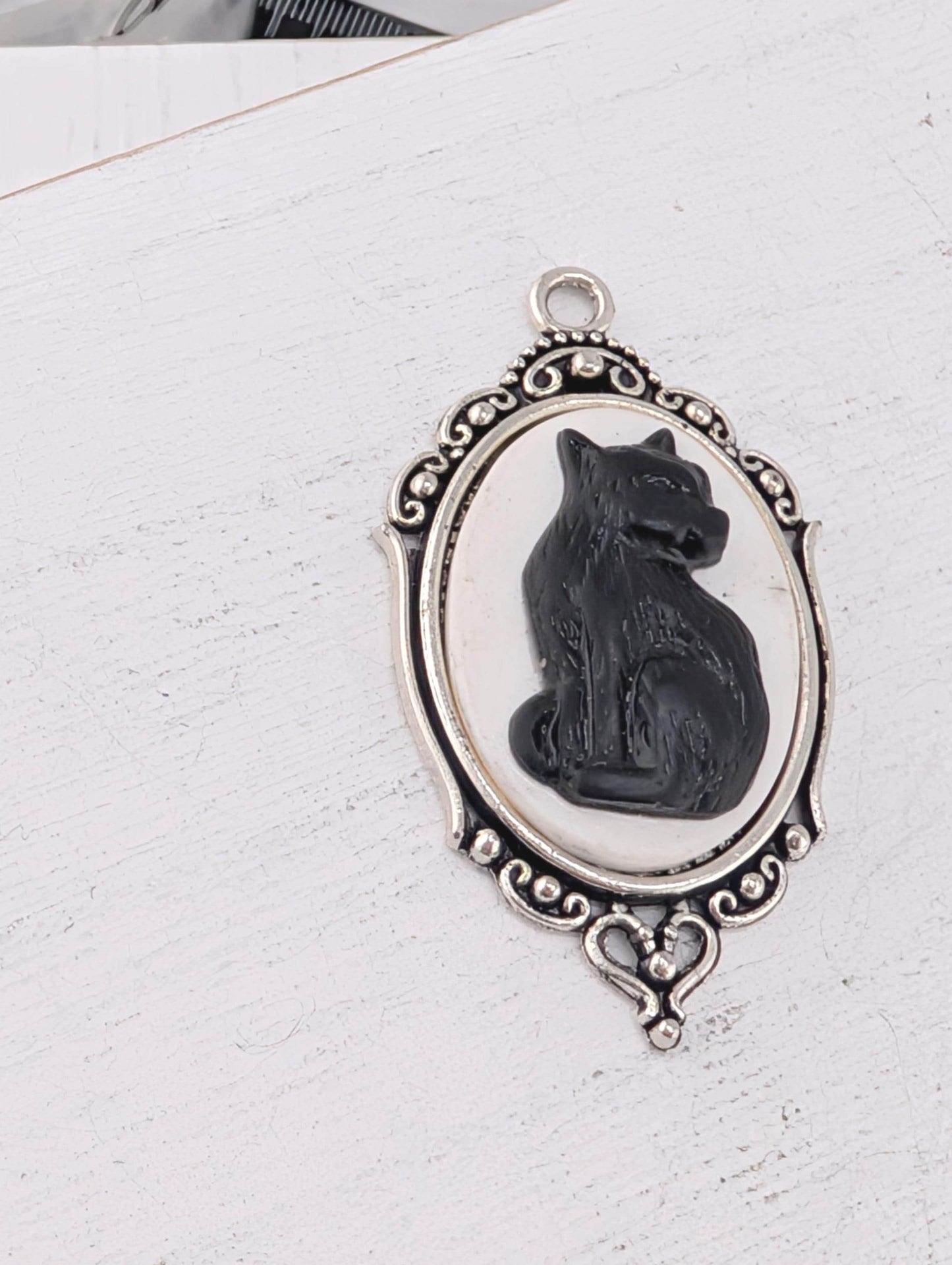A silver-toned pendant featuring a black cat sitting inside an oval frame with an ornate border.