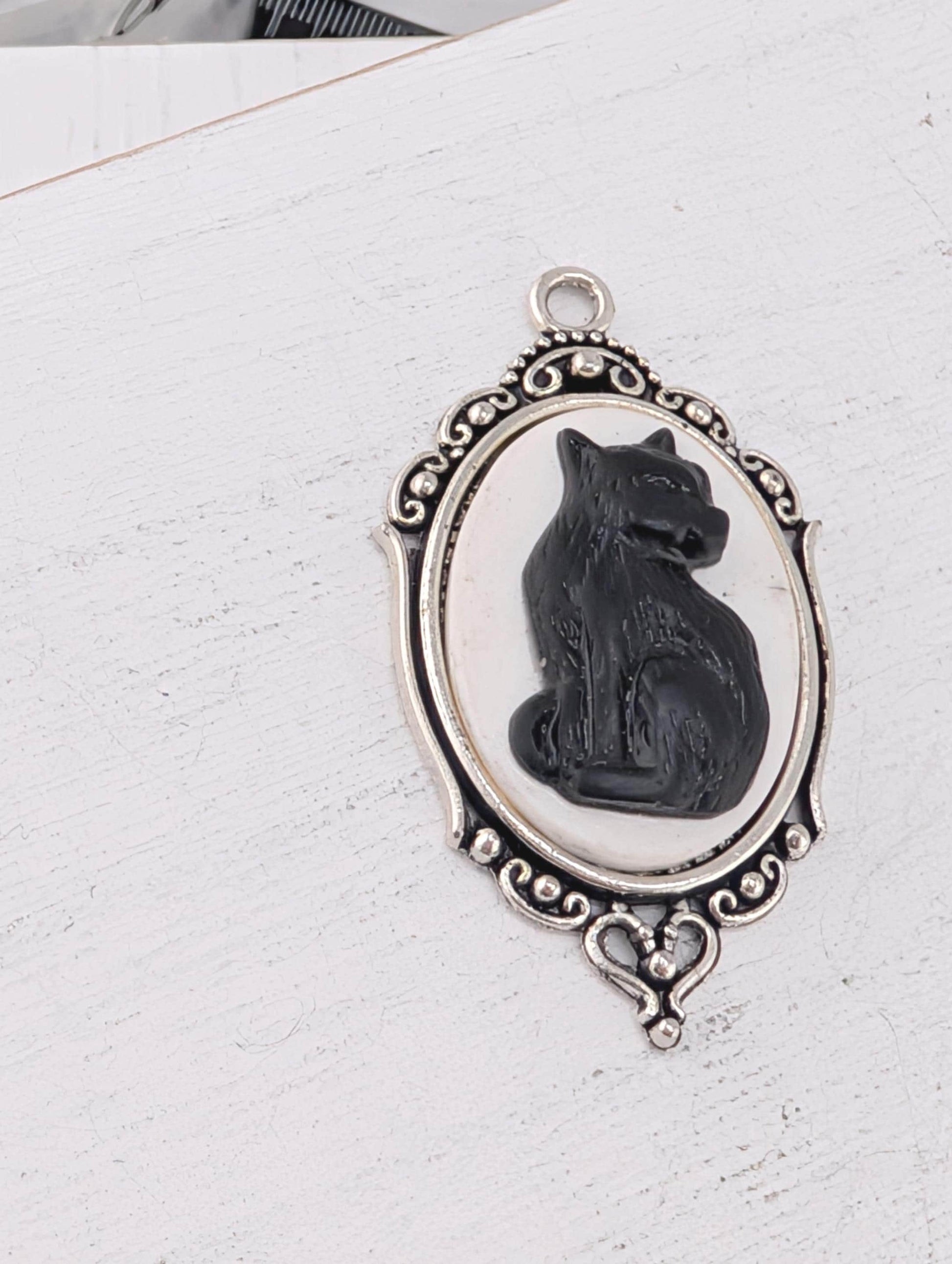 A silver-toned pendant featuring a black cat sitting inside an oval frame with an ornate border.