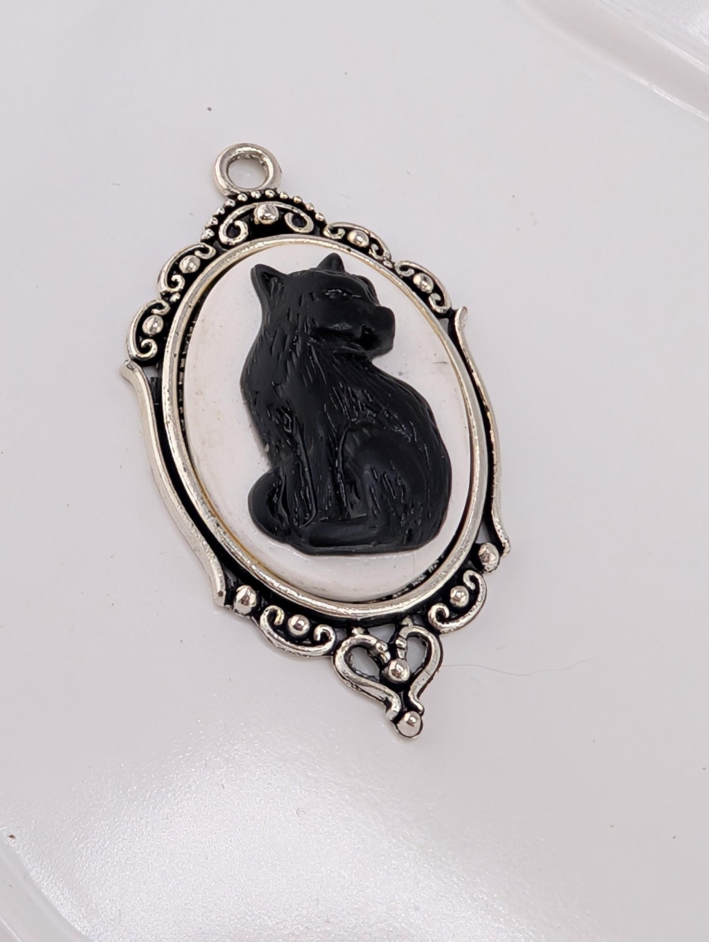 A silver-toned charm in the shape of a cat, with an oval frame and decorative elements, is displayed against a white background.