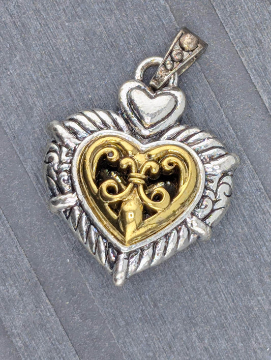 A heart-shaped pendant with a decorative metal design and a small key-like element inside it, resting on a gray surface.
