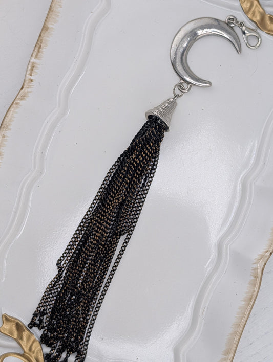 A silver crescent moon pendant with a black tassel hanging from it, displayed on a white surface with a gold-colored frame.