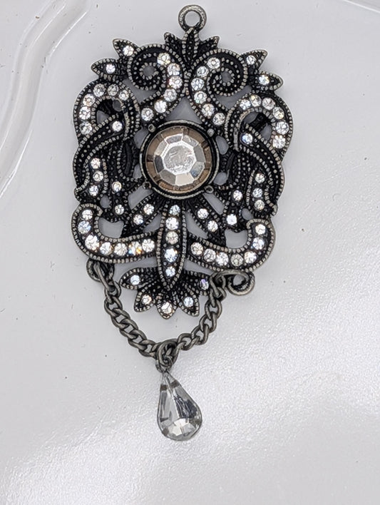 A decorative pendant with a large crystal at the center, surrounded by intricate metalwork and a dangling crystal element.