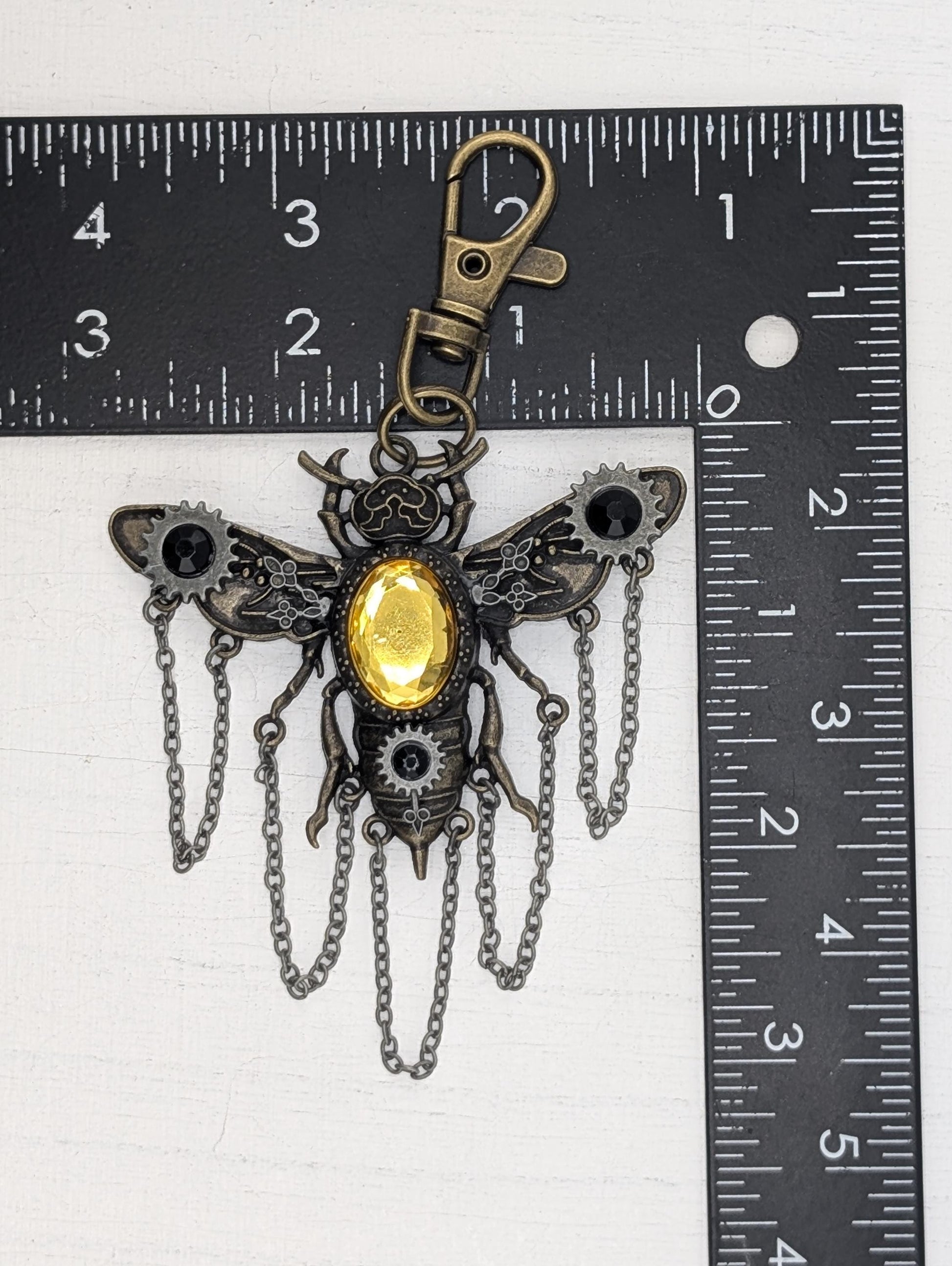 A detailed metal keychain in the shape of a bee, with a yellow gemstone in the center and chains hanging from its wings. The keychain is placed next to a ruler for size comparison.