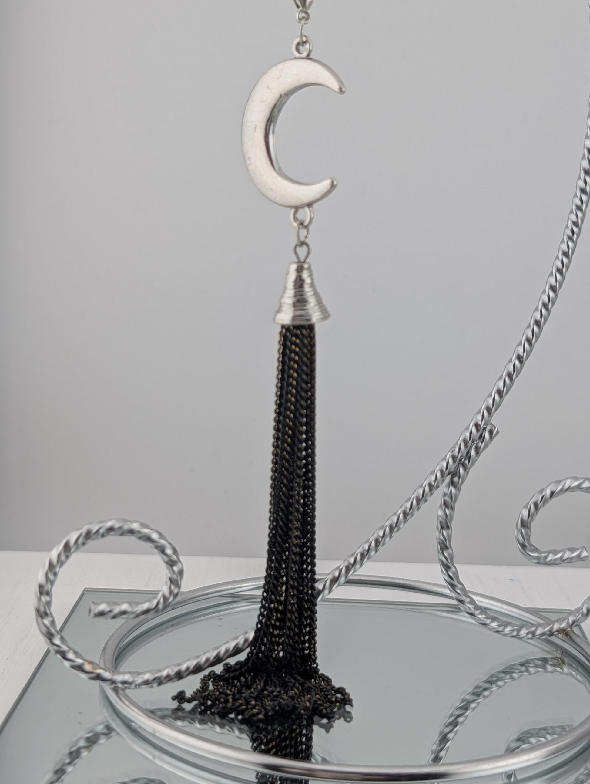 A silver pendant with a crescent moon design and a black tassel hanging from it, displayed on a silver stand.