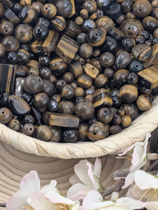 a collection of various beads and stones, including tiger eye beads, arranged in a basket. The basket is placed on a wooden surface, and there are some white flowers visible in the background.