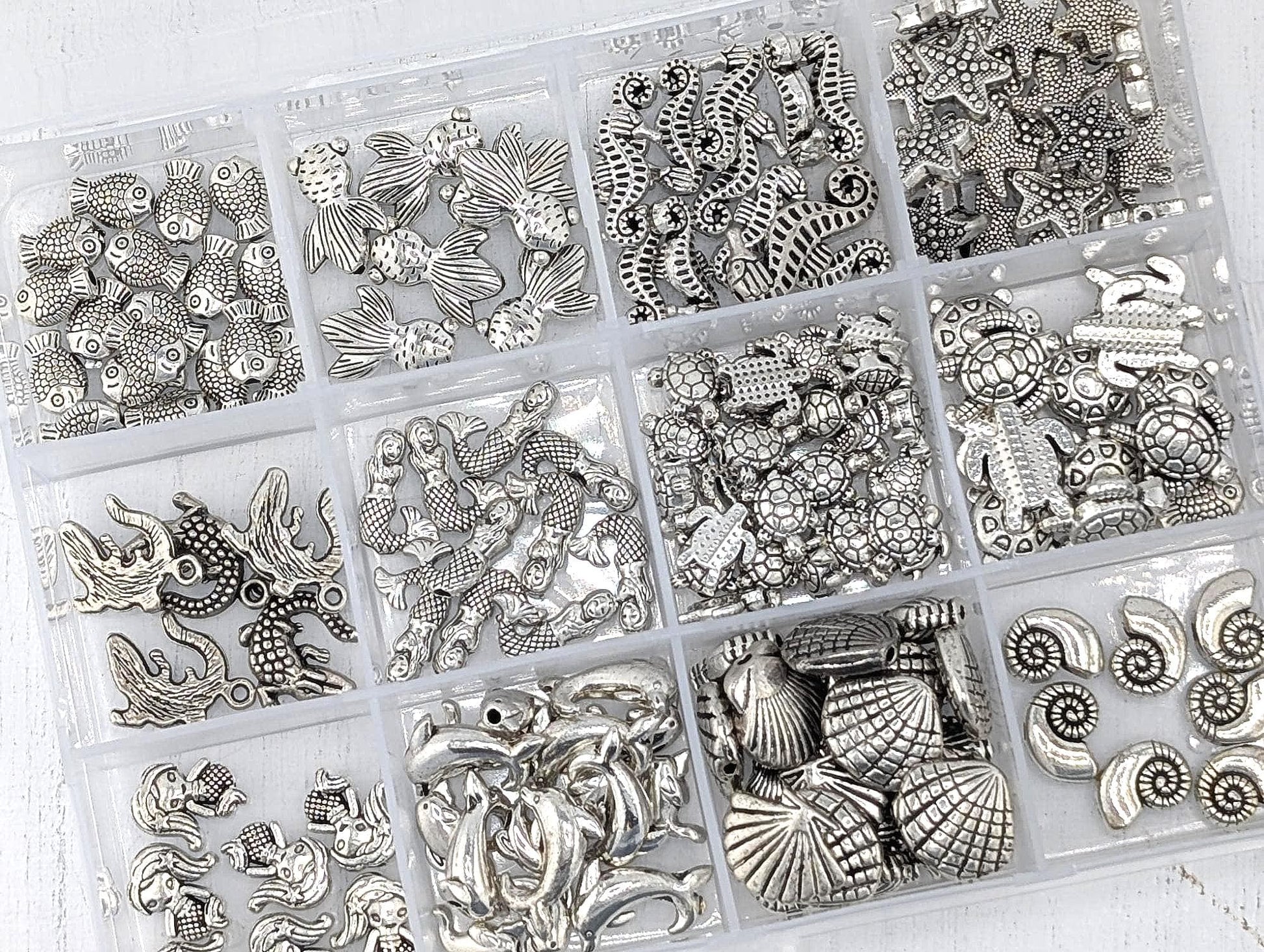 A collection of various silver-colored metal beads and charms, including seashells, starfish, and other marine-themed designs, arranged in multiple compartments.