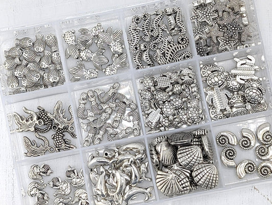 A collection of various silver-colored metal beads and charms, including seashells, starfish, and other marine-themed designs, arranged in multiple compartments.