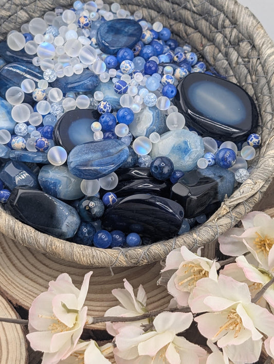 A woven basket filled with various blue and white stones, including crystals and gemstones, alongside delicate white flowers.