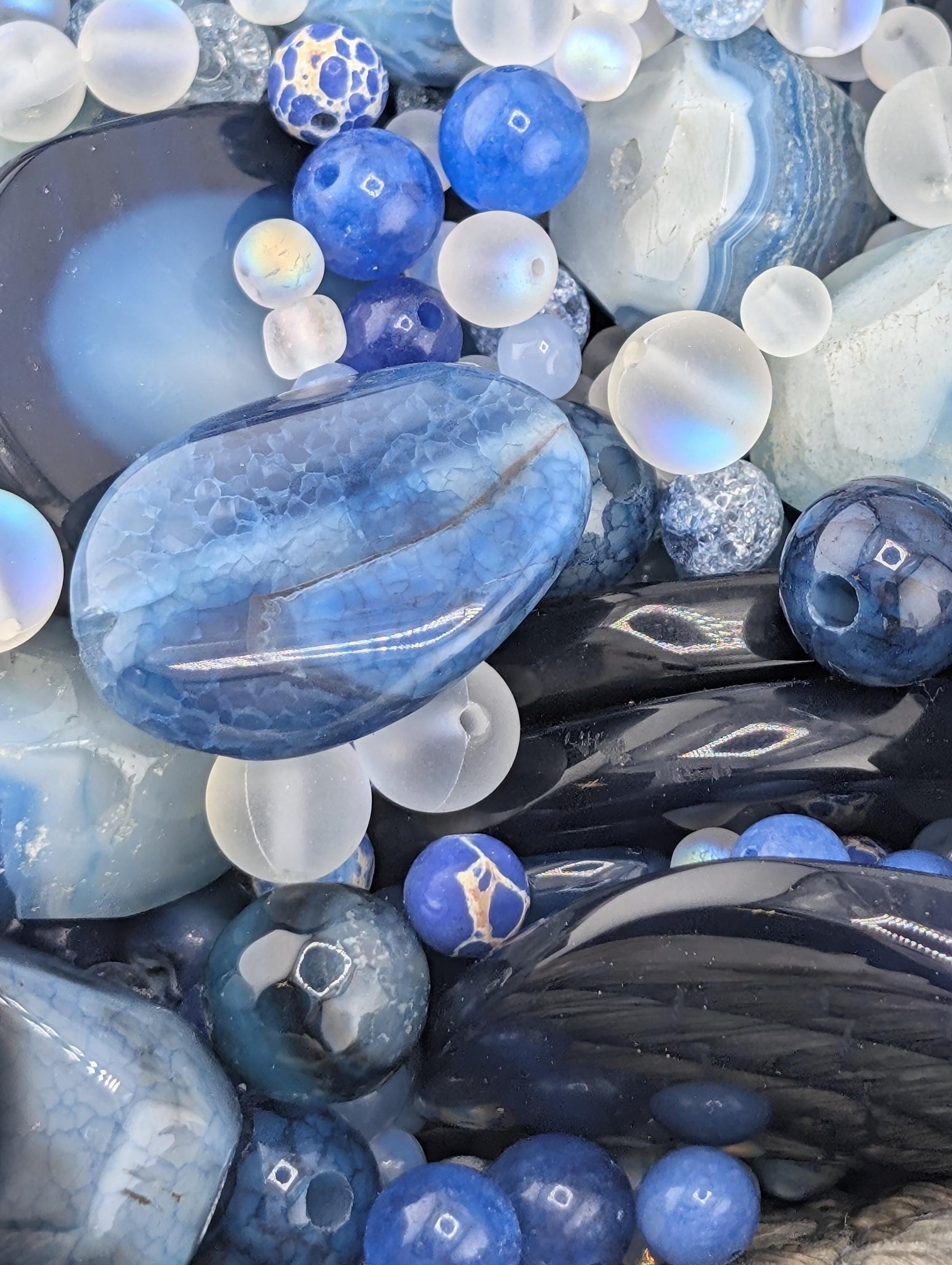 The image is a close-up of various blue and white gemstones and beads, including clear and black stones, as well as multicolored beads.