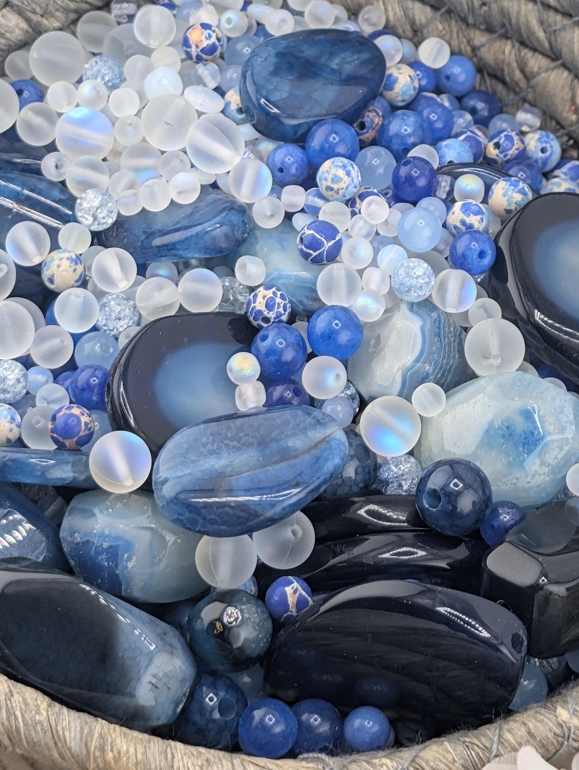 a collection of various blue and white gemstones and beads, including clear and blue stones, as well as some black and white beads, all arranged in a basket.