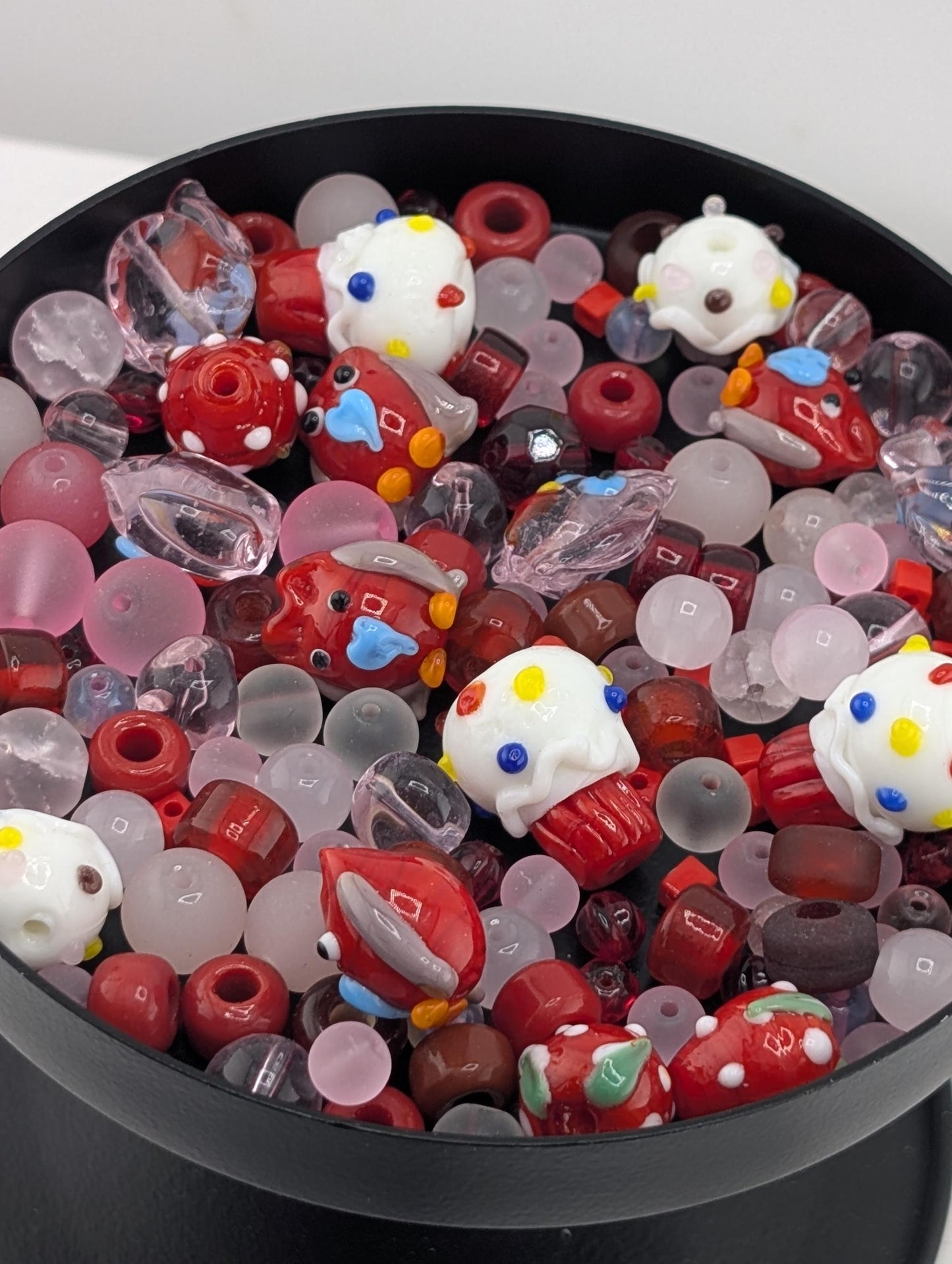 A black bowl filled with a variety of colorful glass beads, including red, white, and multicolored beads, as well as some with polka dots and other playful designs.