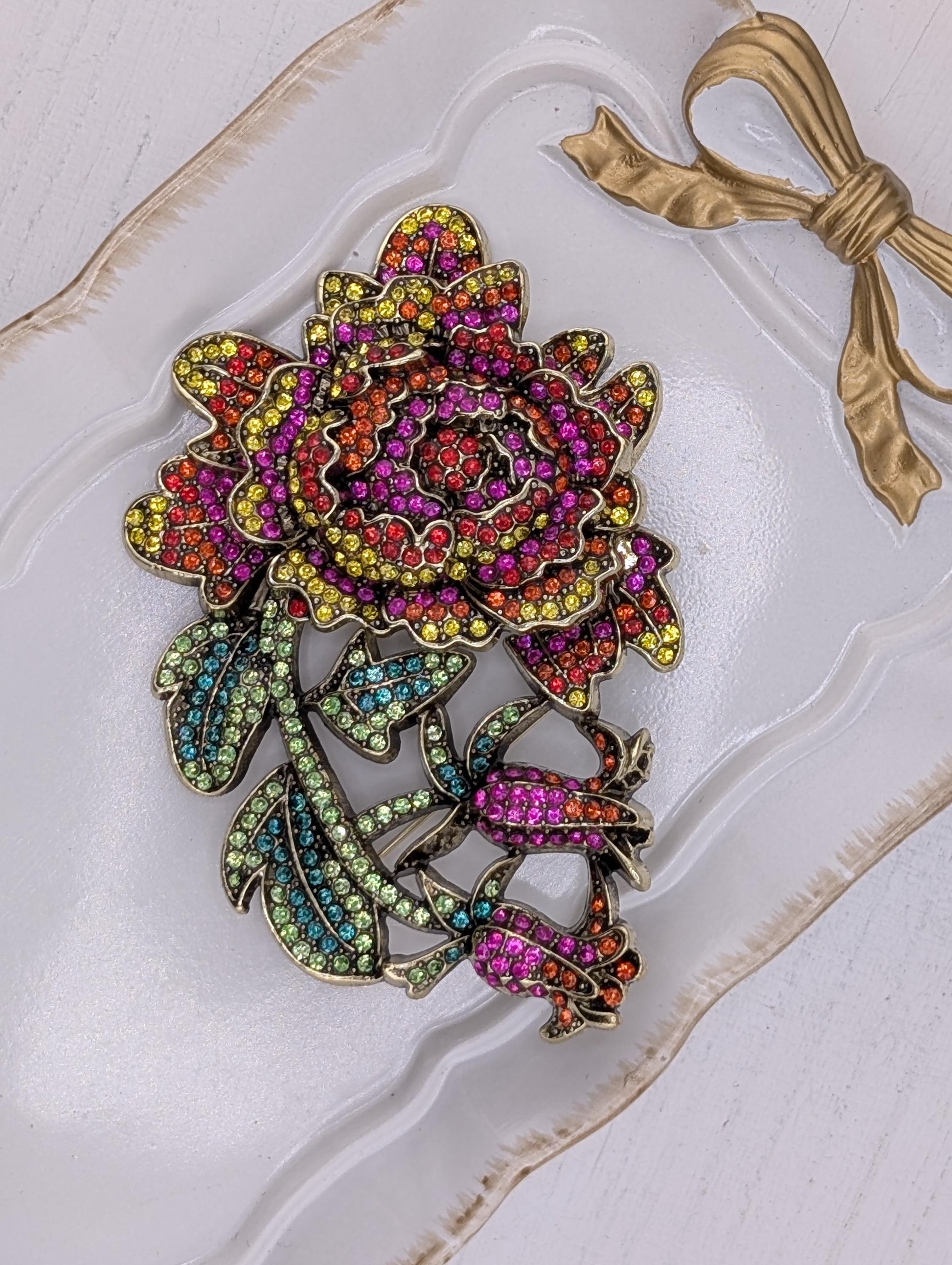 A decorative brooch with a large, colorful flower design and a gold ribbon bow.