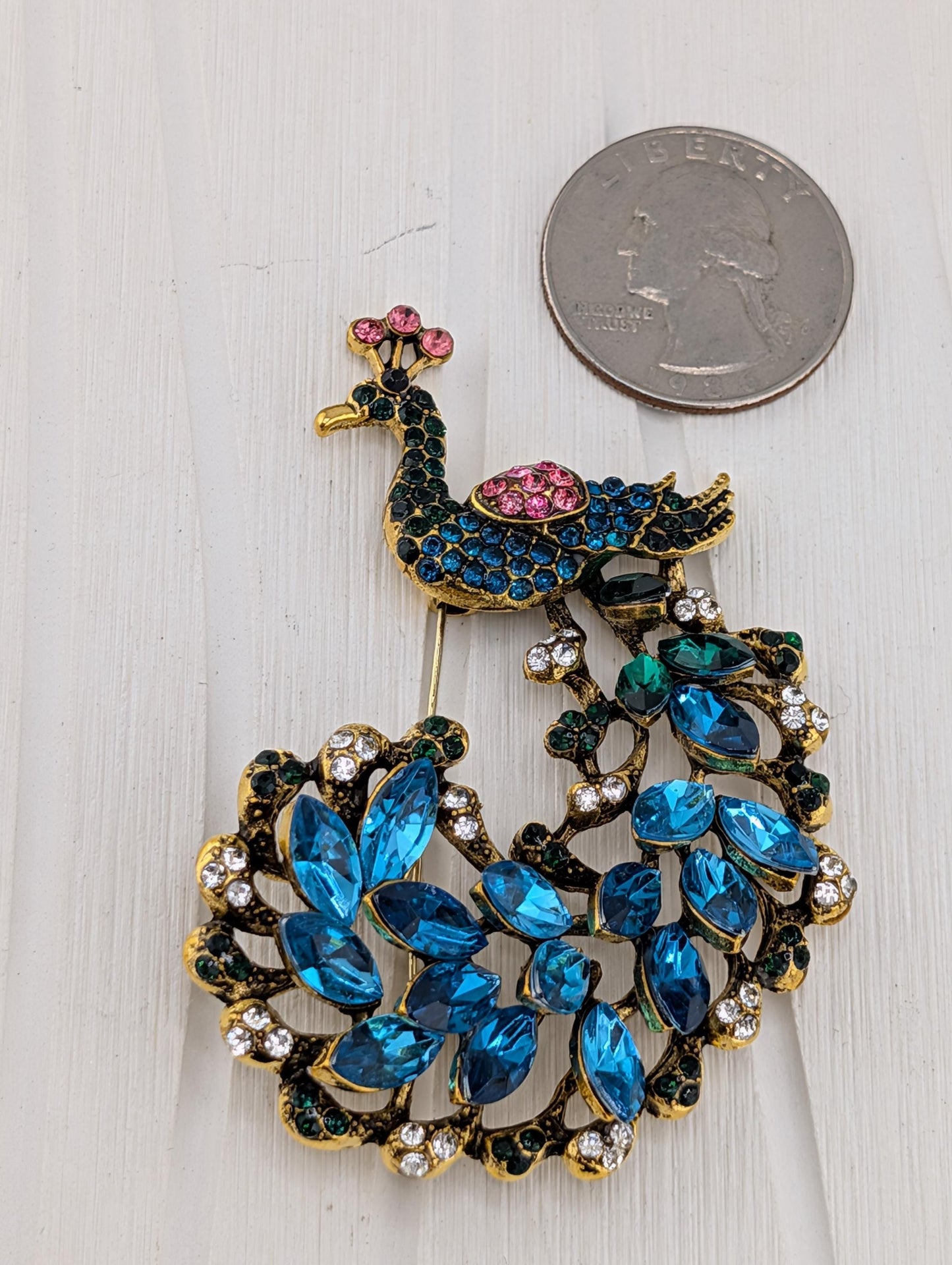 A decorative peacock brooch with blue and green gemstones, placed next to a United States quarter coin.