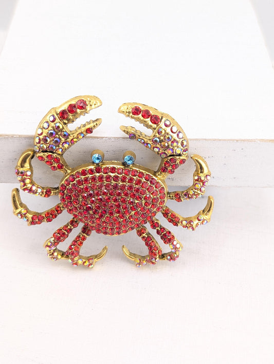 A decorative crab brooch with a red and gold color scheme, adorned with numerous sparkling gemstones.