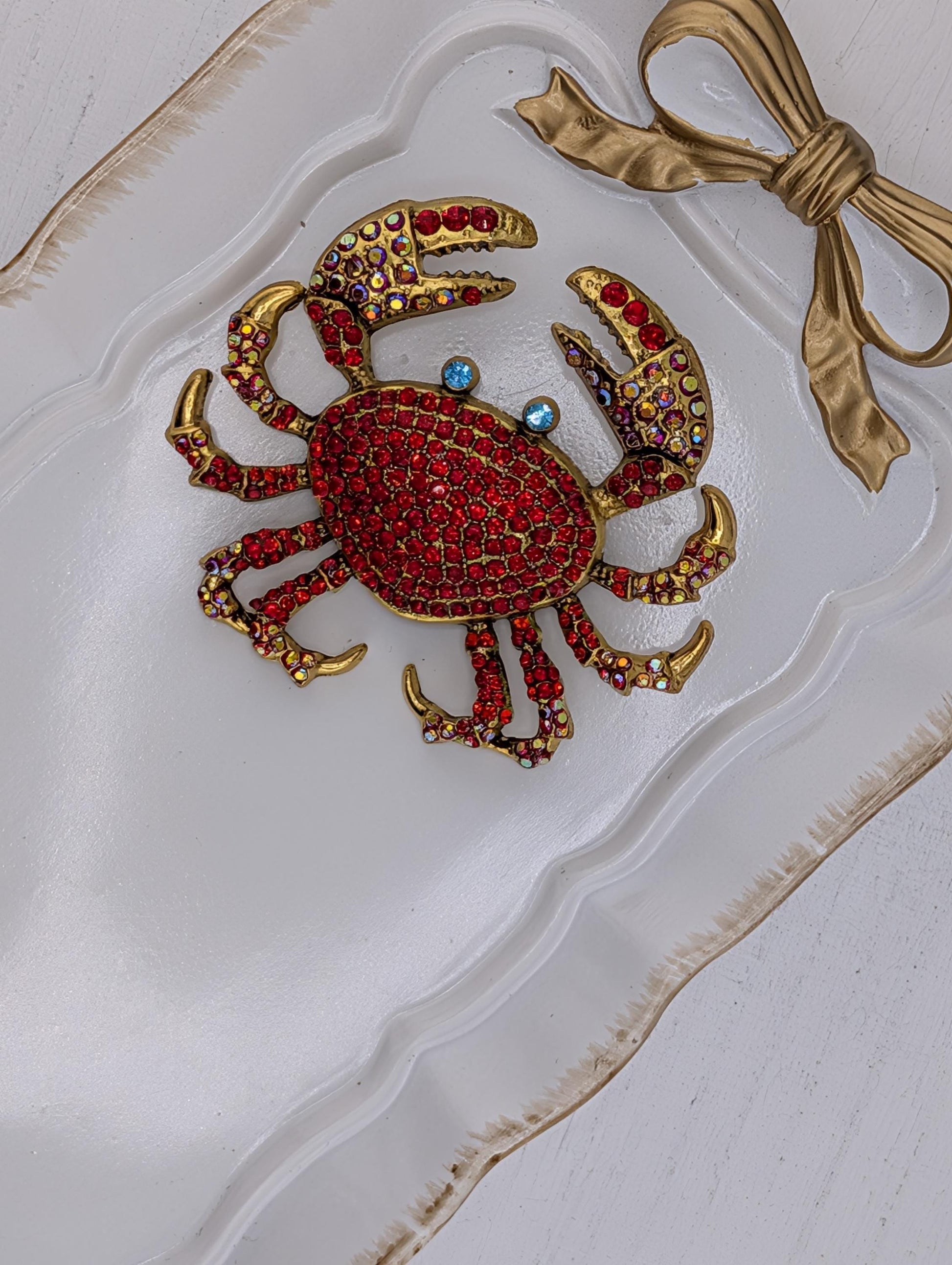 A decorative crab-shaped brooch with red and gold gemstones, placed on a white surface with a gold ribbon bow.