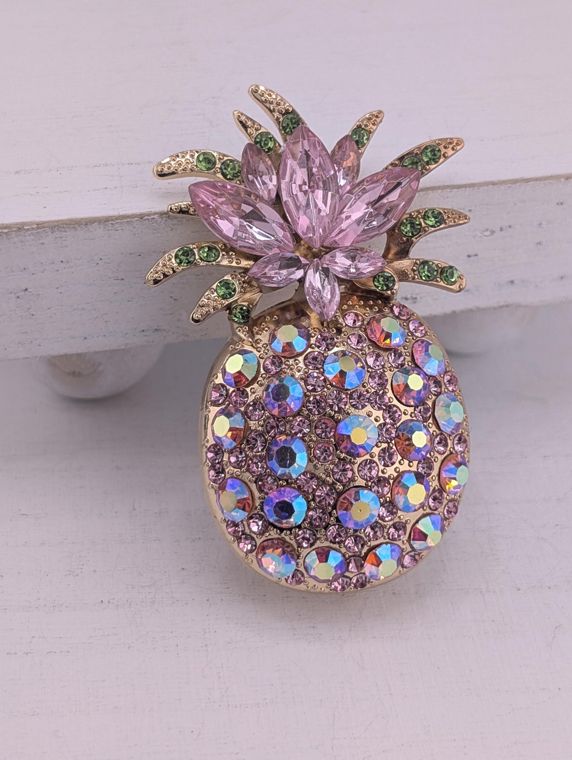 A decorative pin or brooch featuring a pineapple design with pink and purple gemstones, including a large flower-like element and smaller circular stones.